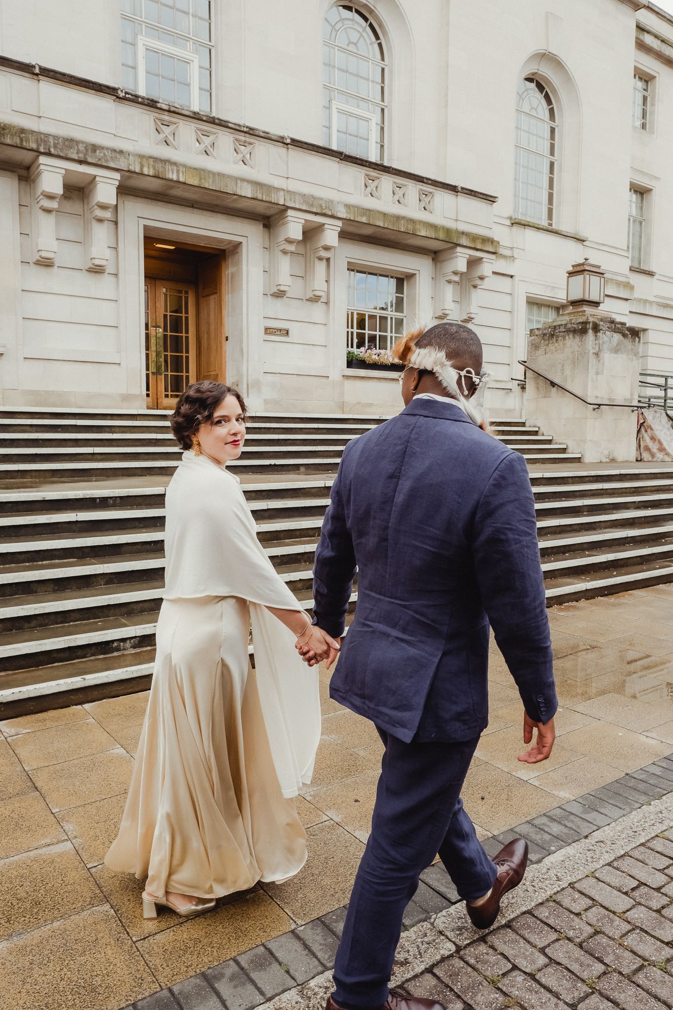 A couple in formal attire ascends stone steps outside a historic building on a rainy day.