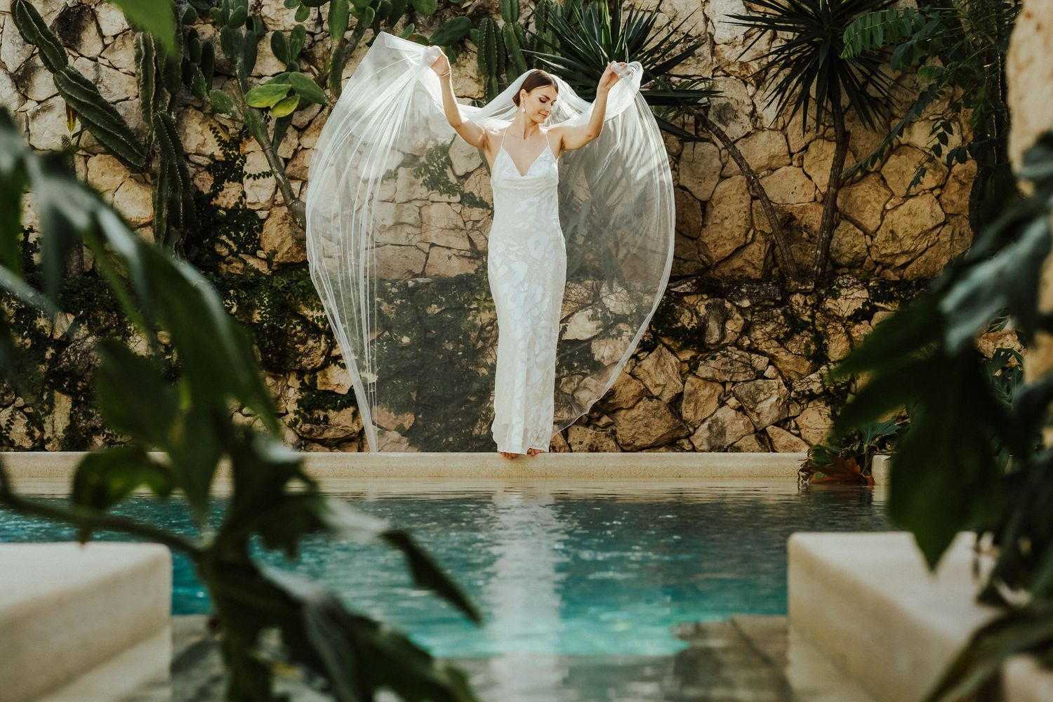 Elegant bride in white gown poses by stone wall and pool surrounded by tropical greenery.