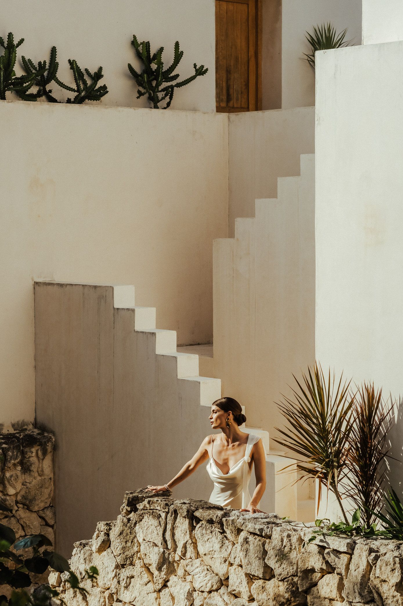 A sequence of four photos showing a person in white dress exploring a rustic stone wall with interior stairs and plants.
