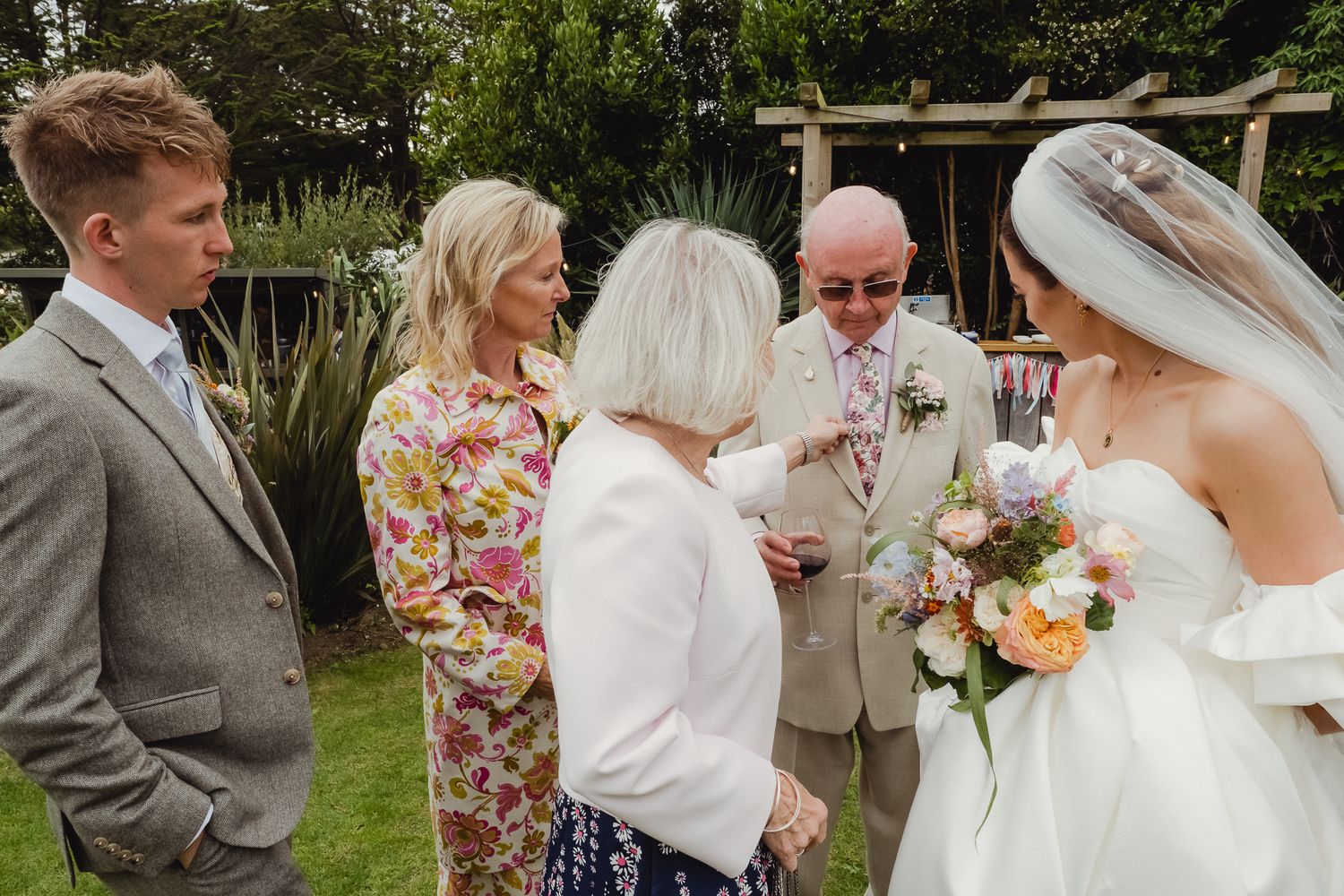 A small group gathers outdoors in a garden setting during a wedding ceremony on a sunny day.