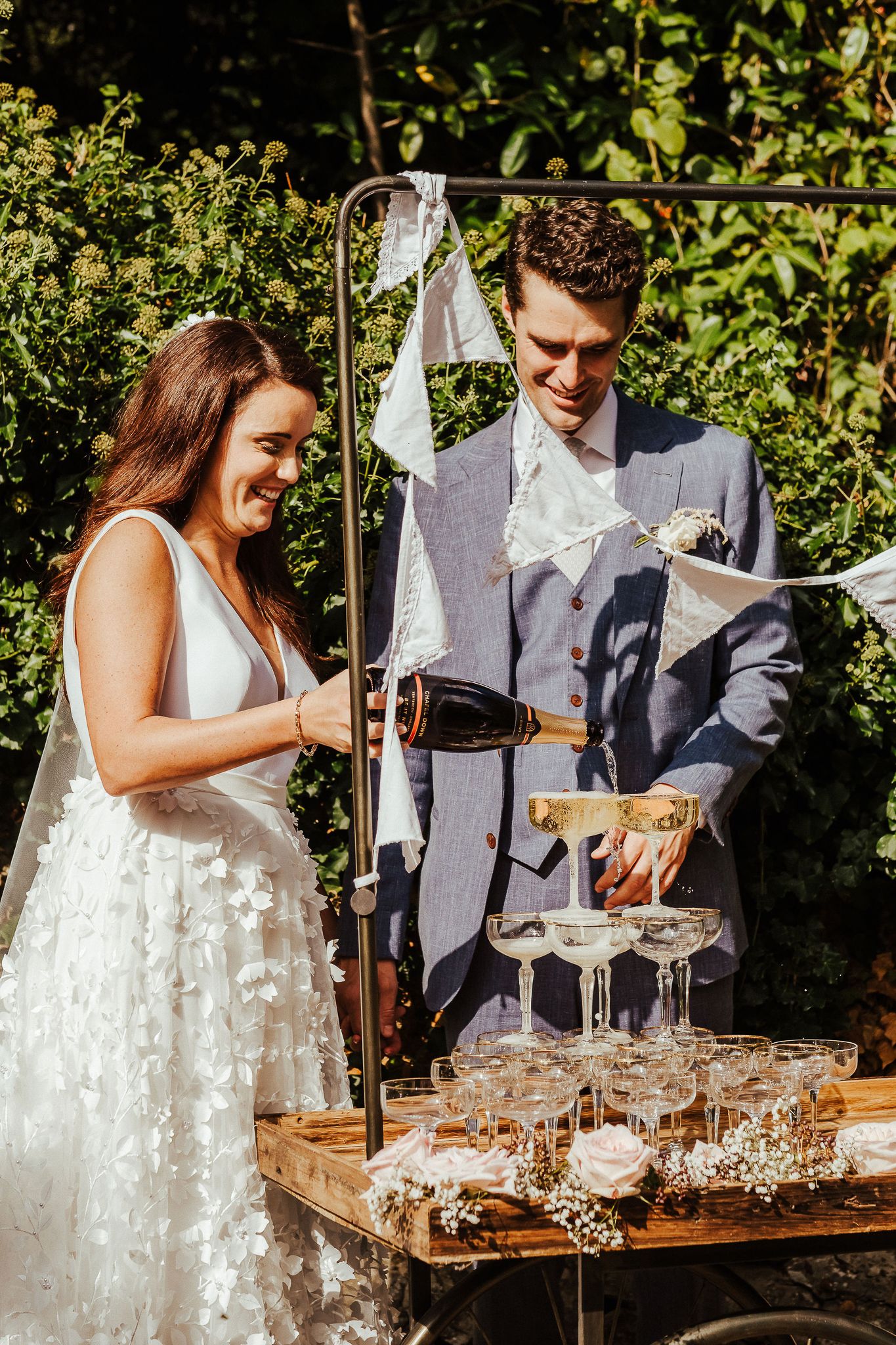 A couple pouring champagne into a tower of glasses at their outdoor wedding reception.