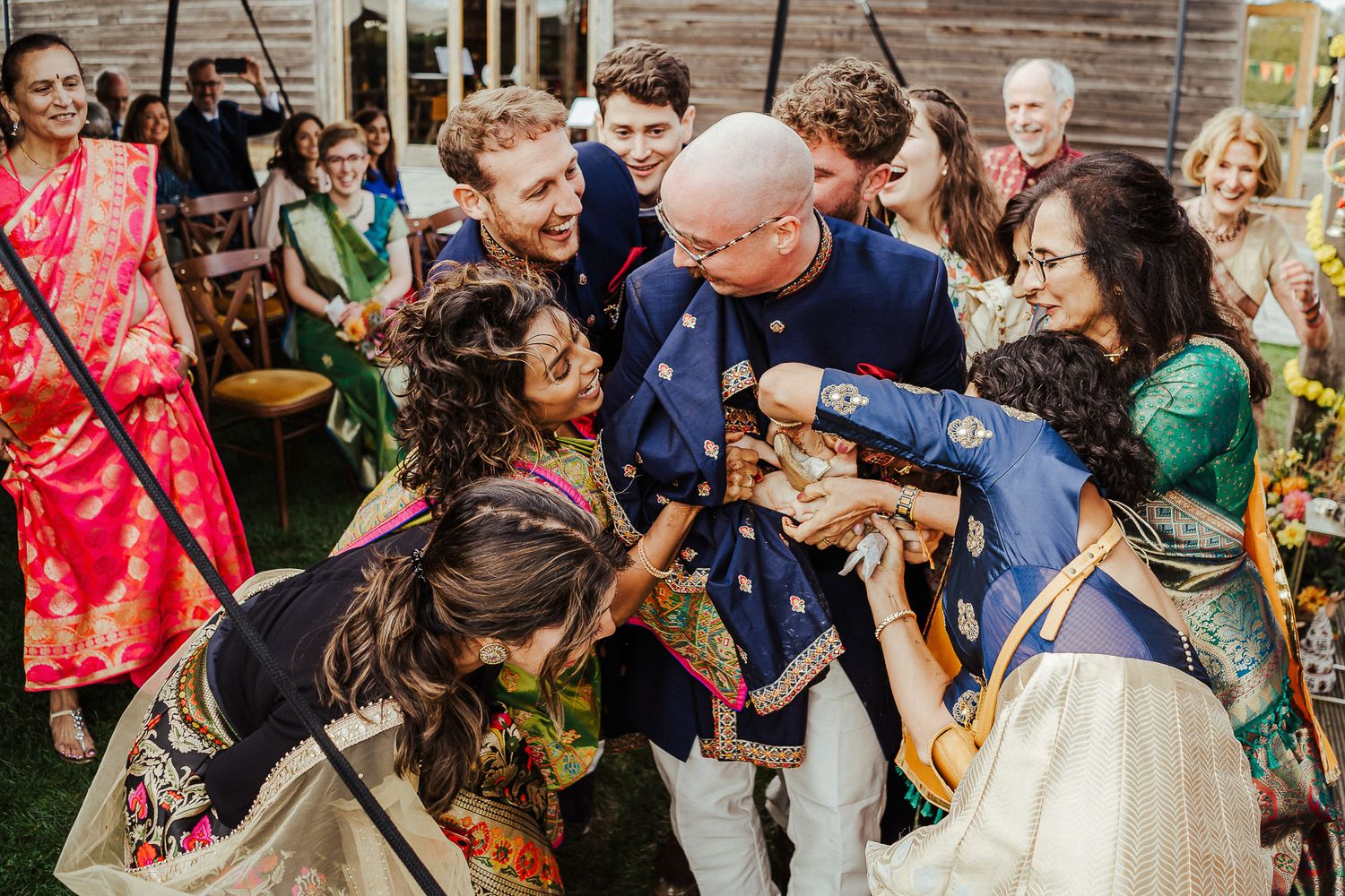 A joyful group celebrates at an outdoor wedding reception wearing traditional Indian attire.