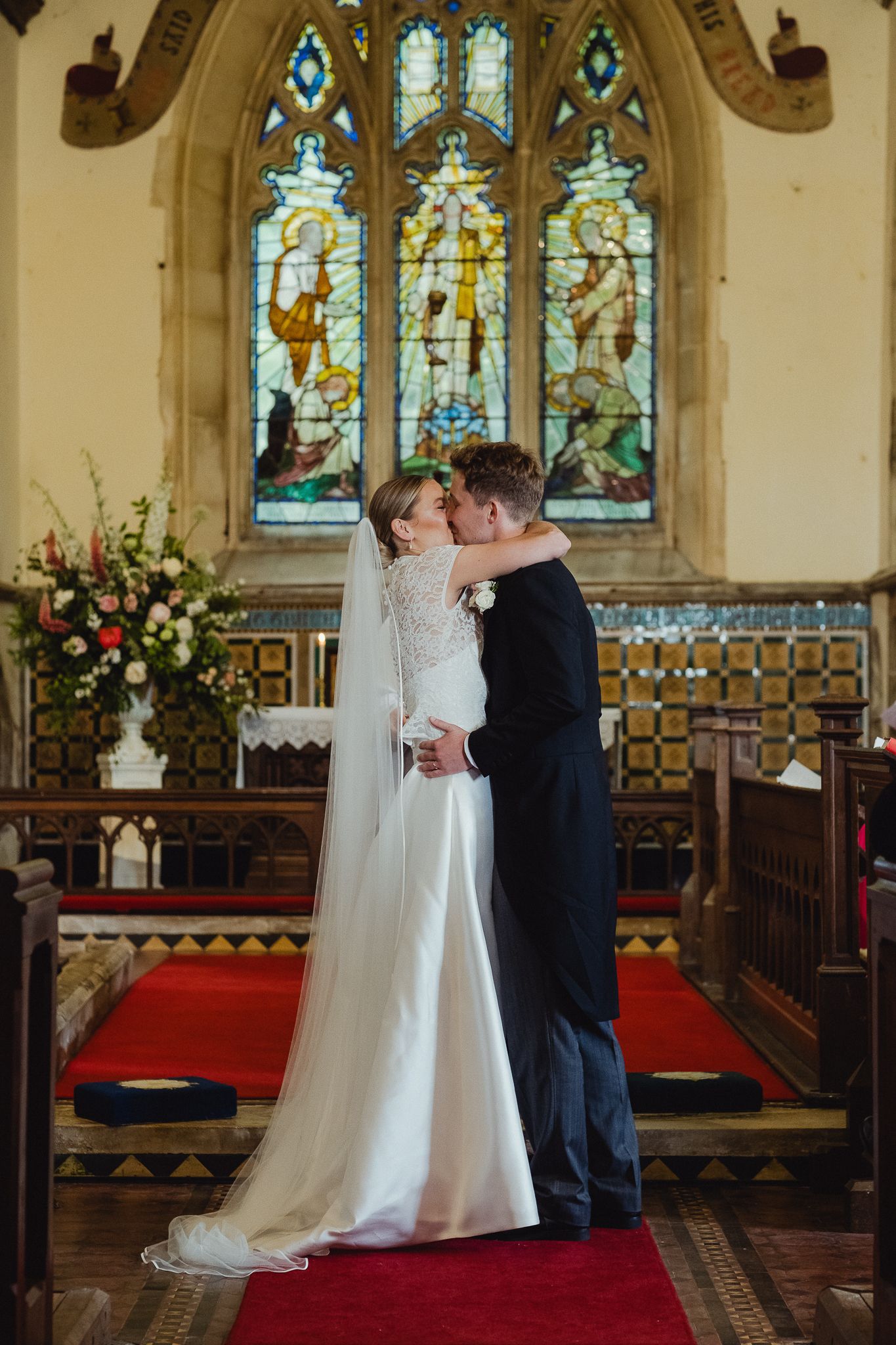 A romantic wedding ceremony in a church with stained glass windows, red carpet aisle, and floral arrangements.