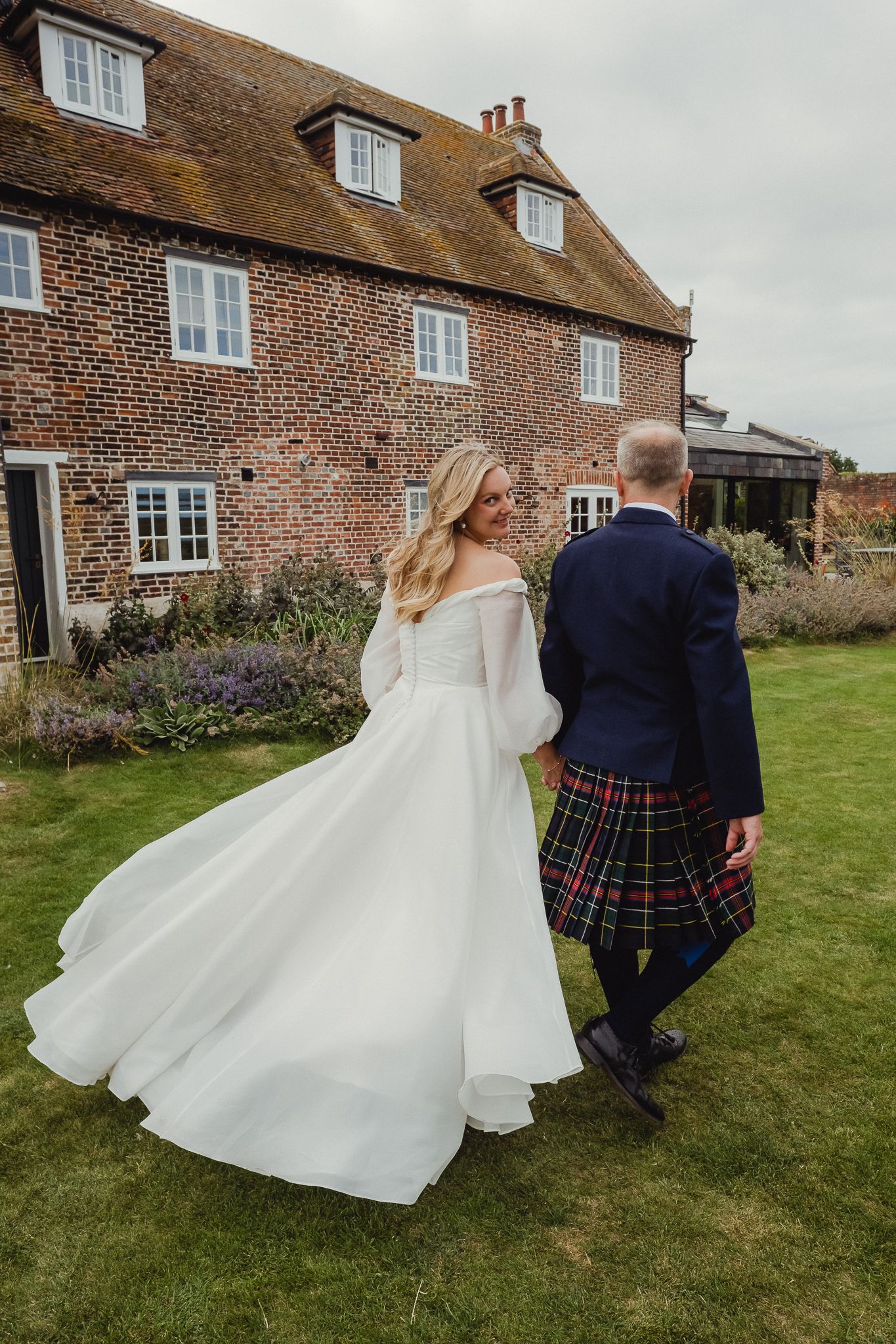 A wedding couple stands on a lawn outside a historic brick farmhouse with a thatched roof and dormer windows.
