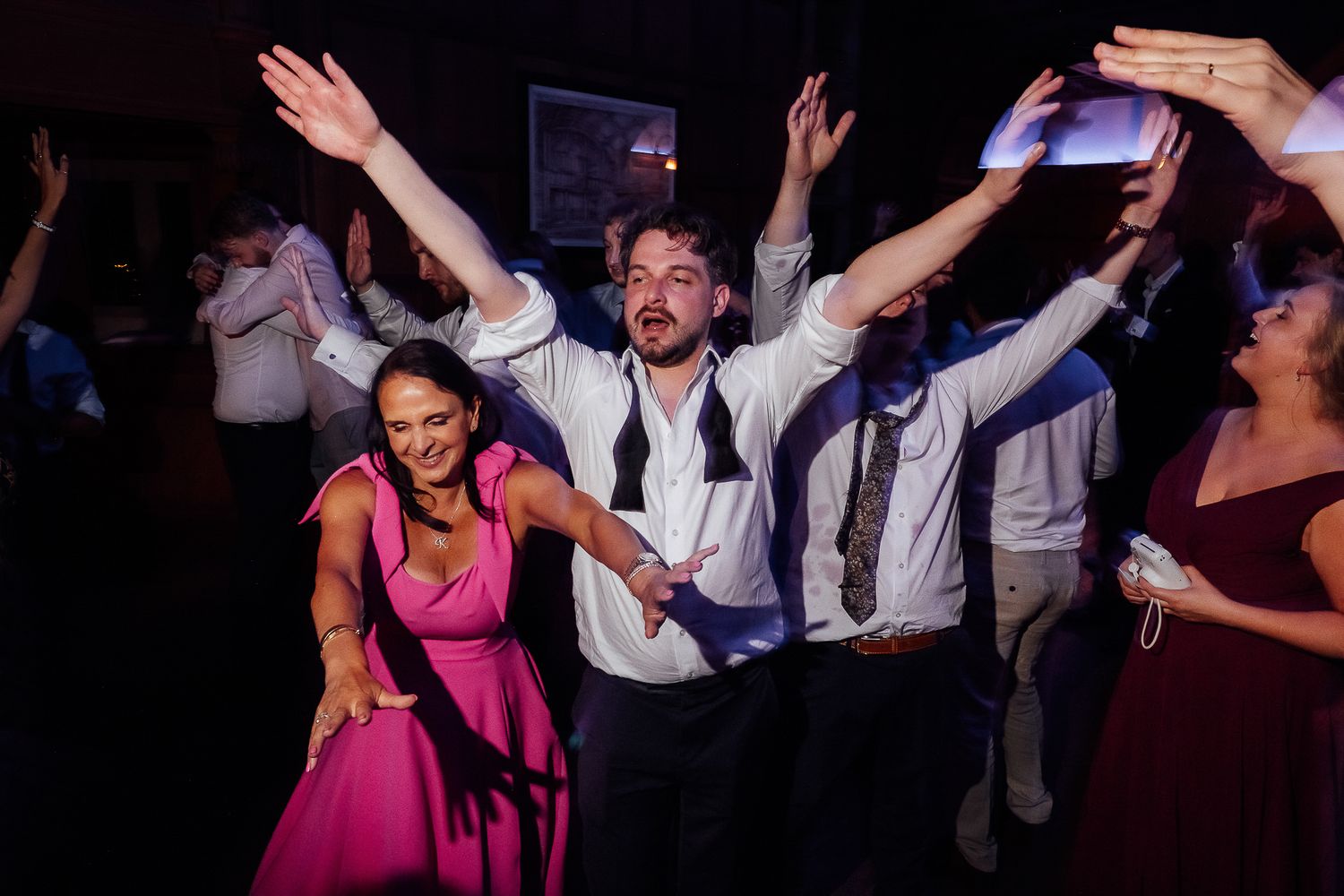 Couple dancing joyfully at a wedding reception, woman in pink dress and man with tie loosened, hands raised.