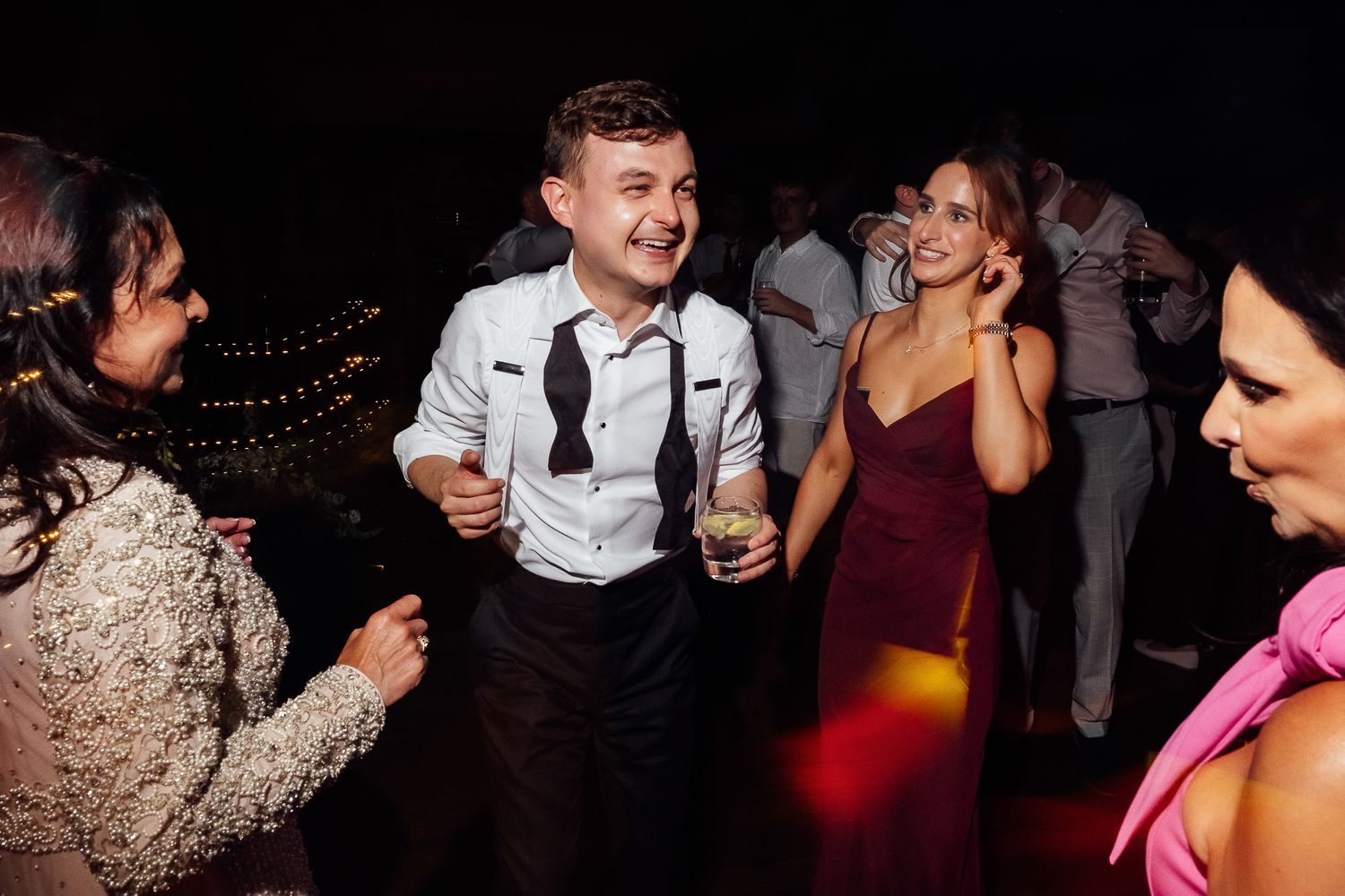 Young man in suspenders dancing joyfully at a wedding reception with guests in formal attire.