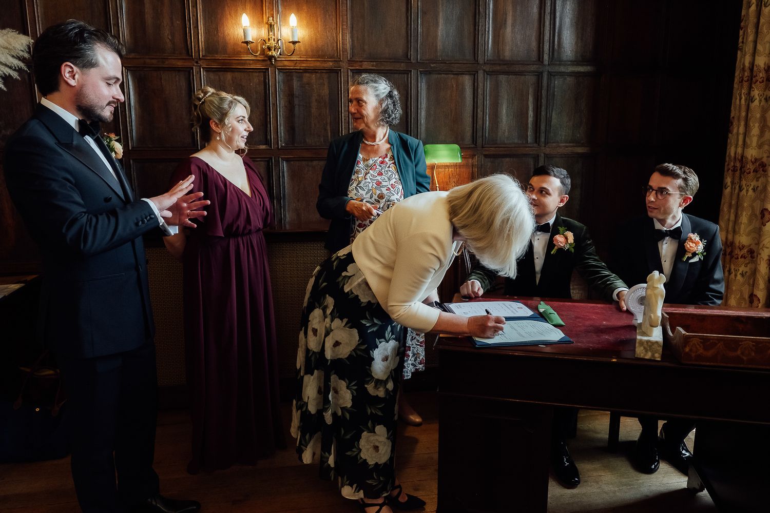 Wedding signing ceremony in wood-paneled room with guests watching as officiant signs marriage register at antique desk.