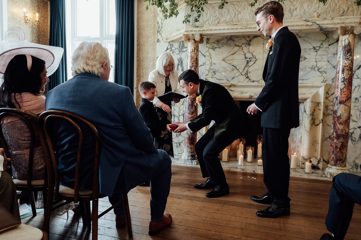 Groomsmen in black suits gather near a marble fireplace during a wedding ceremony in an elegant venue.