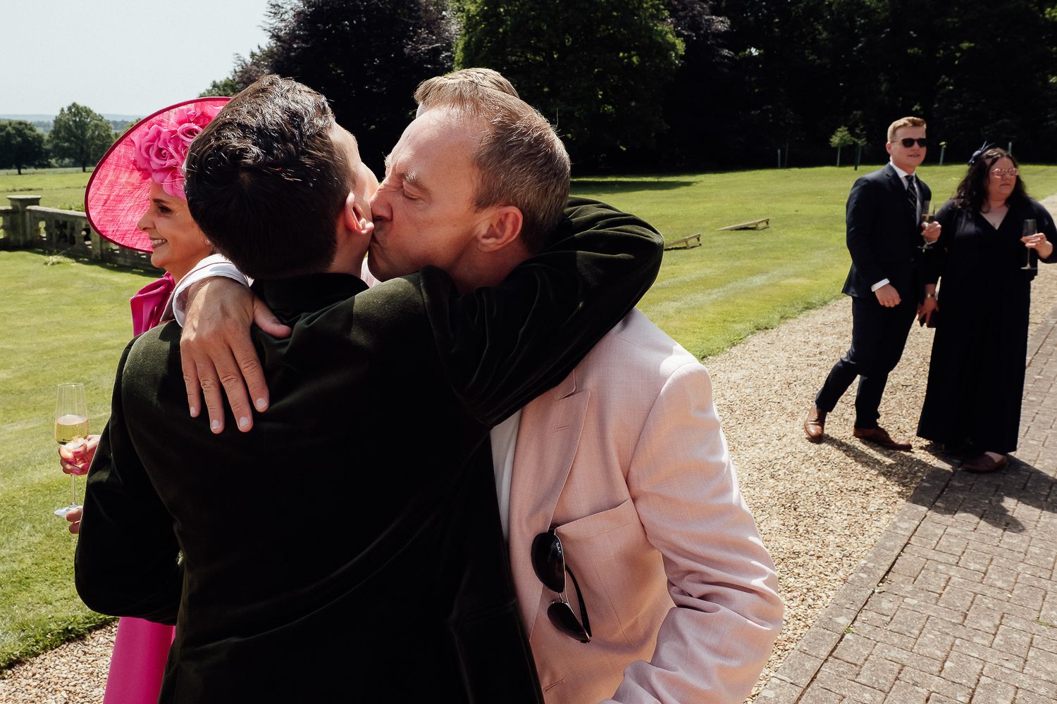 Two men share a kiss at an outdoor wedding celebration, with guests in formal attire visible in the background.