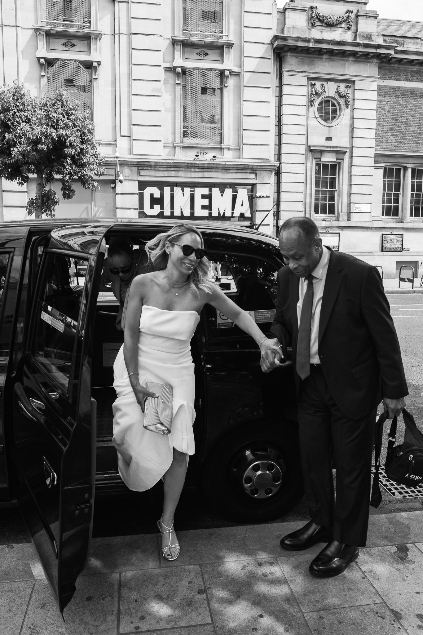 Smiling bride in a white strapless dress and sunglasses steps out of a black taxi, helped by a man in a suit outside a cinema.