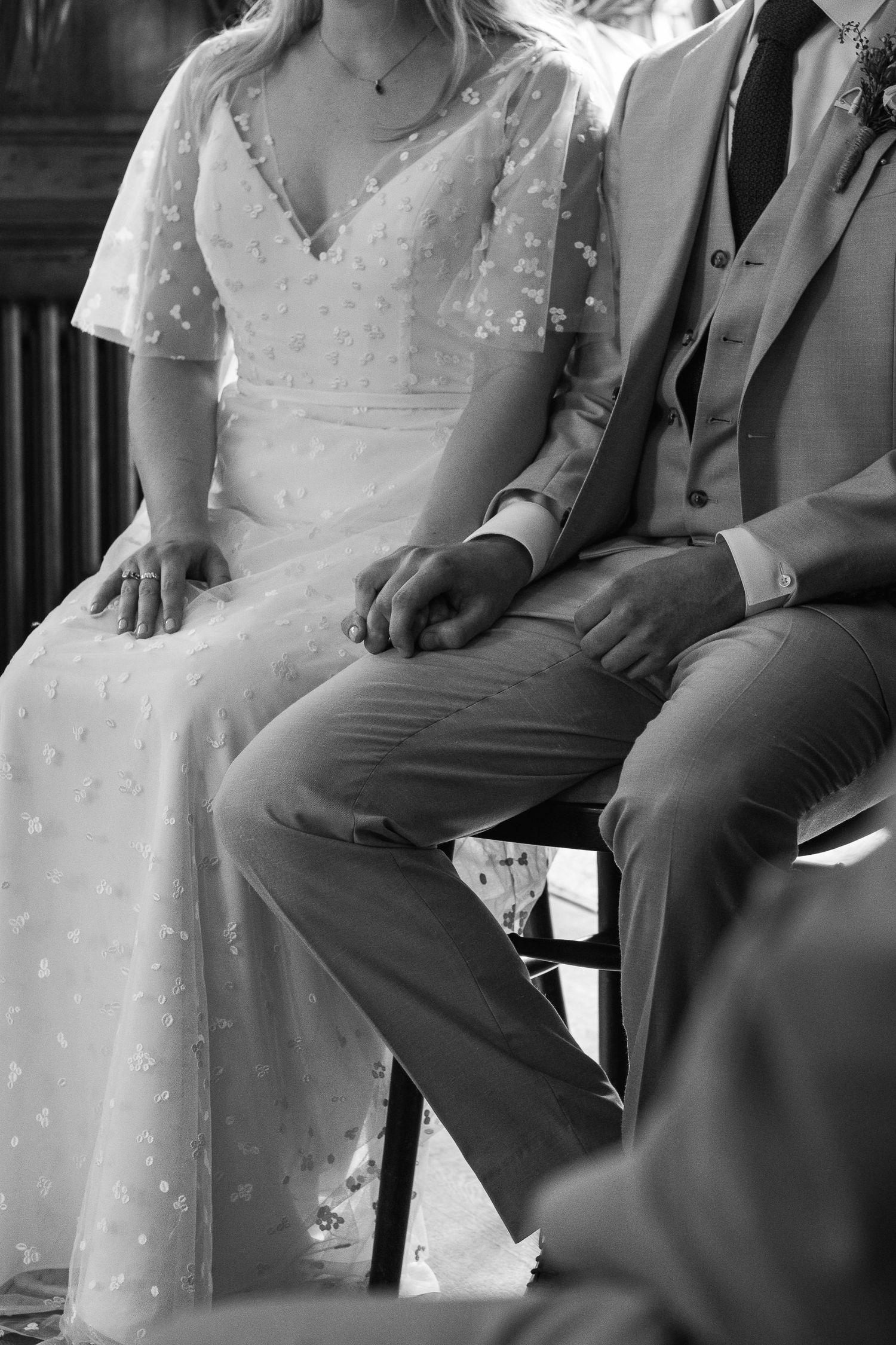 Black and white photo of a seated bride in a floral lace wedding dress holding hands with a groom in a three-piece suit.