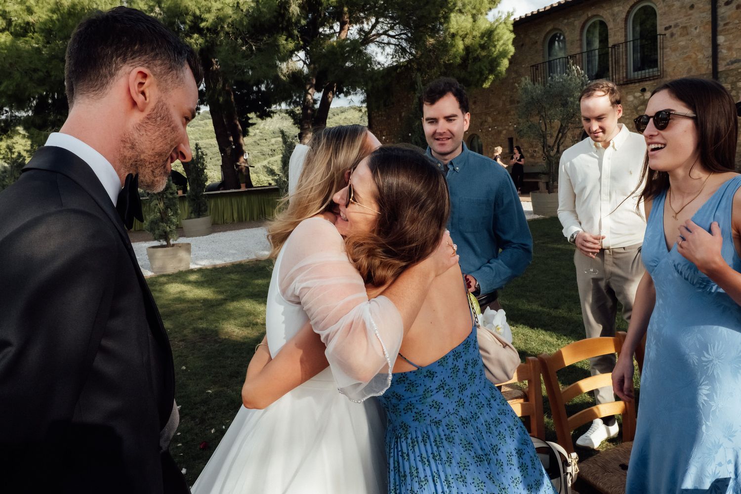 A bride in a white gown embraces a woman in a blue dress at an outdoor wedding while smiling guests look on.