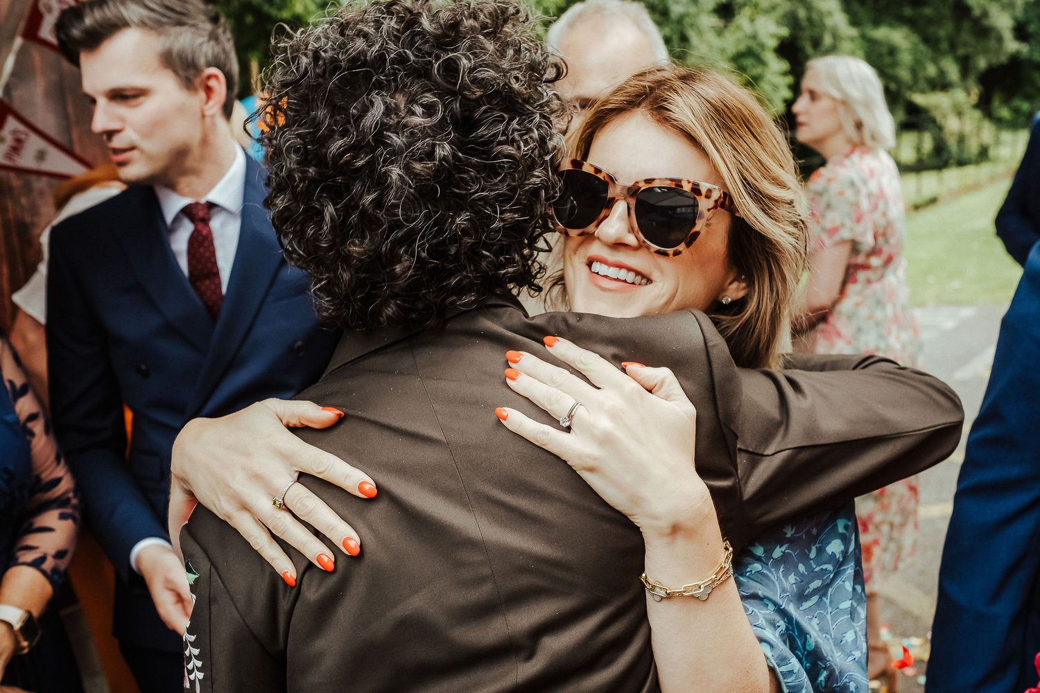 Smiling woman in sunglasses with orange manicured nails hugs a curly-haired person at an outdoor gathering with guests in the background.