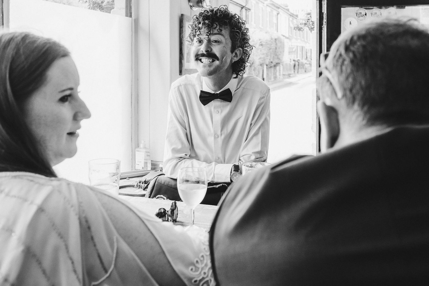 Smiling curly-haired waiter in a bow tie stands at a restaurant table while two seated diners chat in this black and white photo.