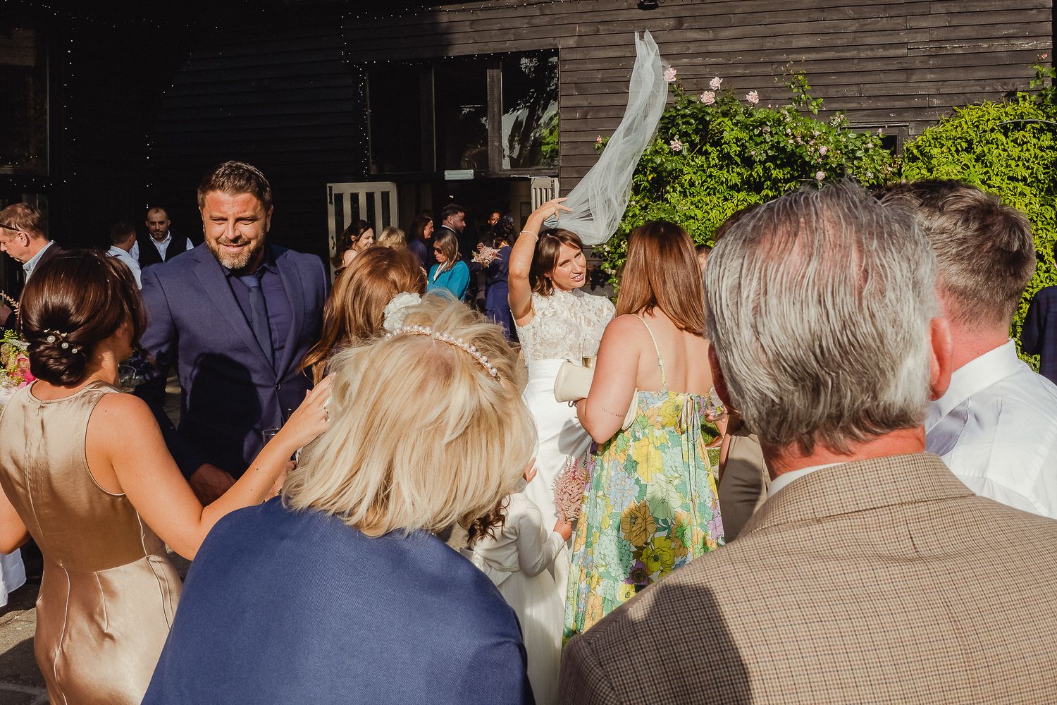 Wedding guests gathered outdoors at a reception venue surrounded by lush greenery and natural lighting.