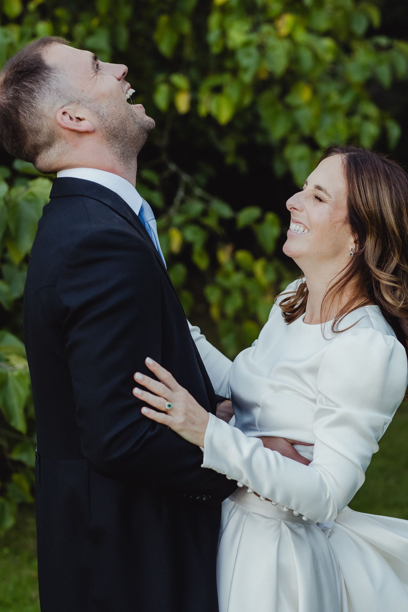 A couple in formal attire share a joyful dance moment outdoors with greenery in the background.