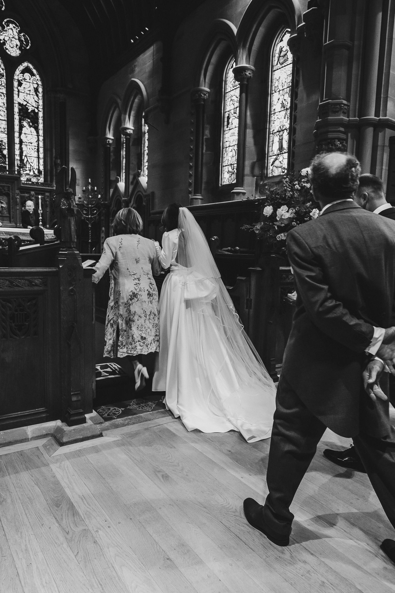 Black and white photo of a bride in a flowing white gown with long veil entering a church with stained glass windows.