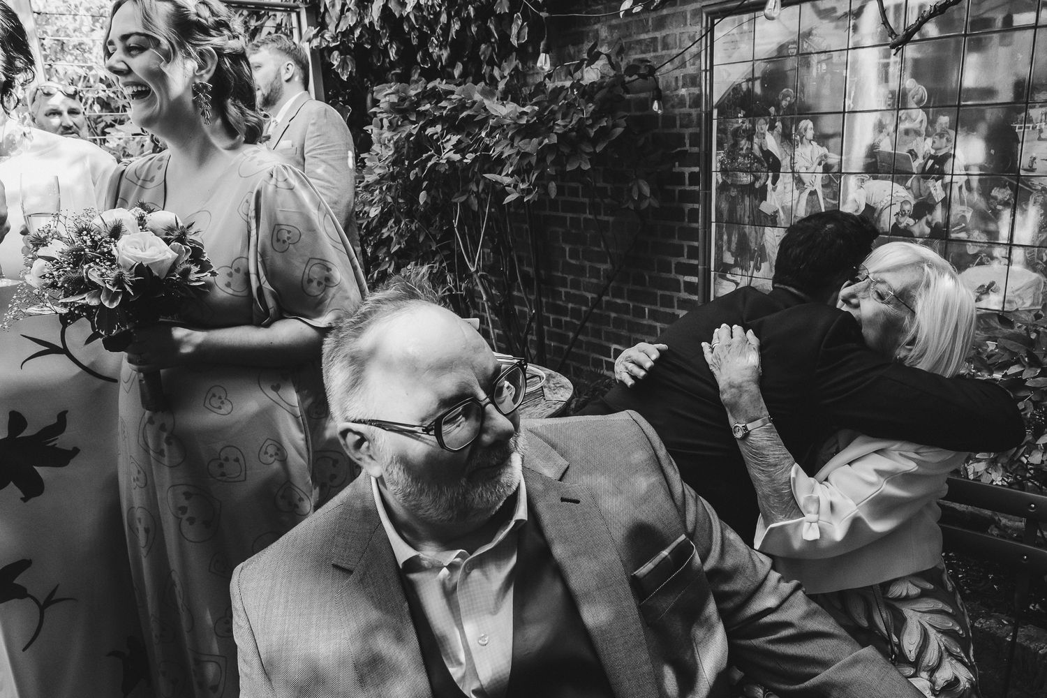 A candid black and white photo captures guests chatting and laughing during an outdoor wedding reception.
