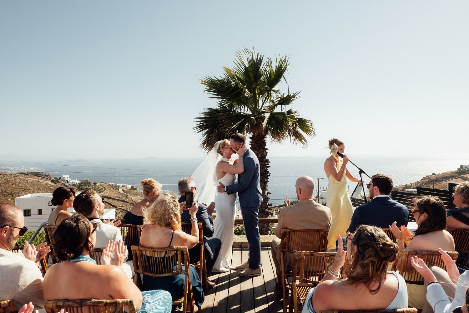 Outdoor wedding ceremony with couple kissing at altar, palm tree, ocean view, guests seated, musician performing on sunny day.