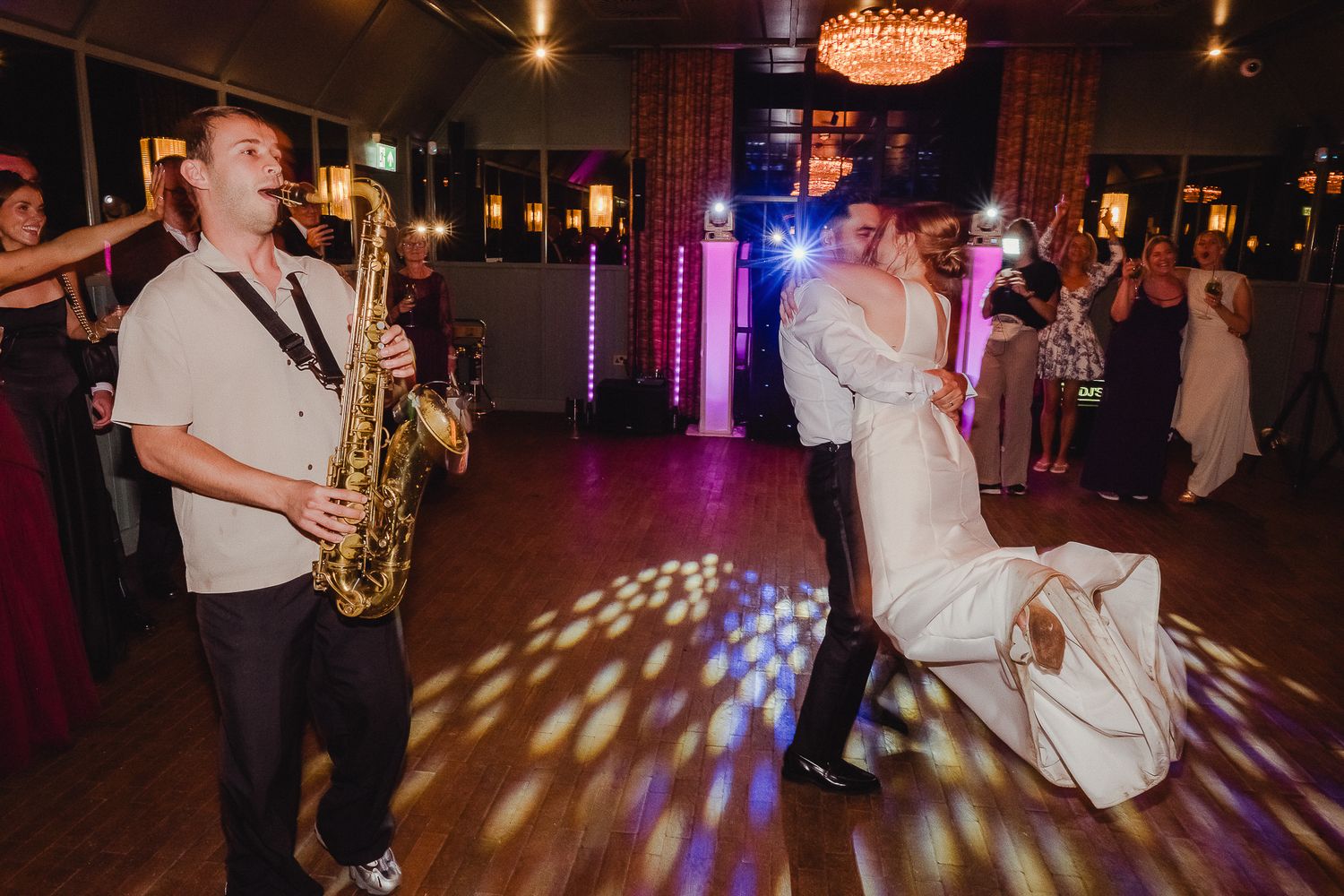 People dancing on a dark dance floor with colorful disco lights and a saxophone player performing at a wedding reception.