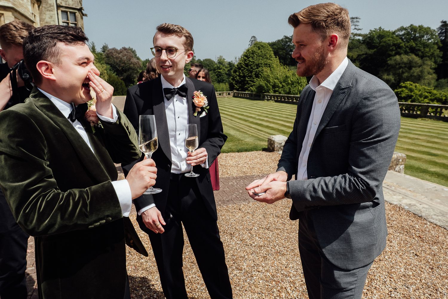 Three men in formal suits socializing outdoors at a wedding reception, holding champagne glasses on a sunny day.