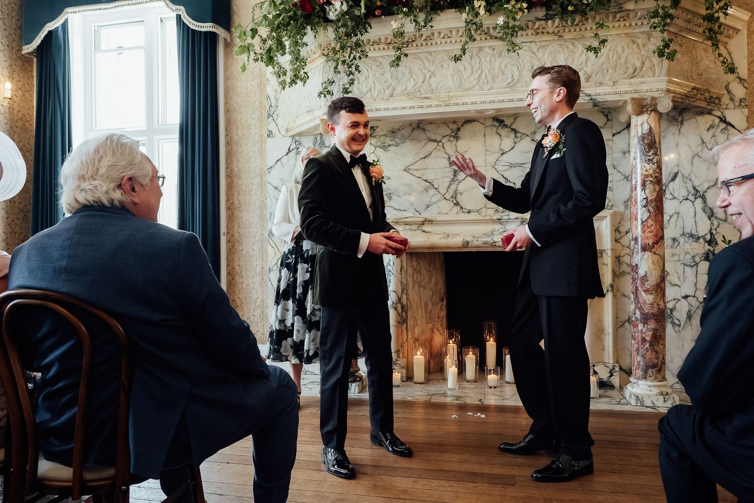 Two grooms exchange vows at an elegant indoor wedding ceremony beside an ornate marble fireplace.