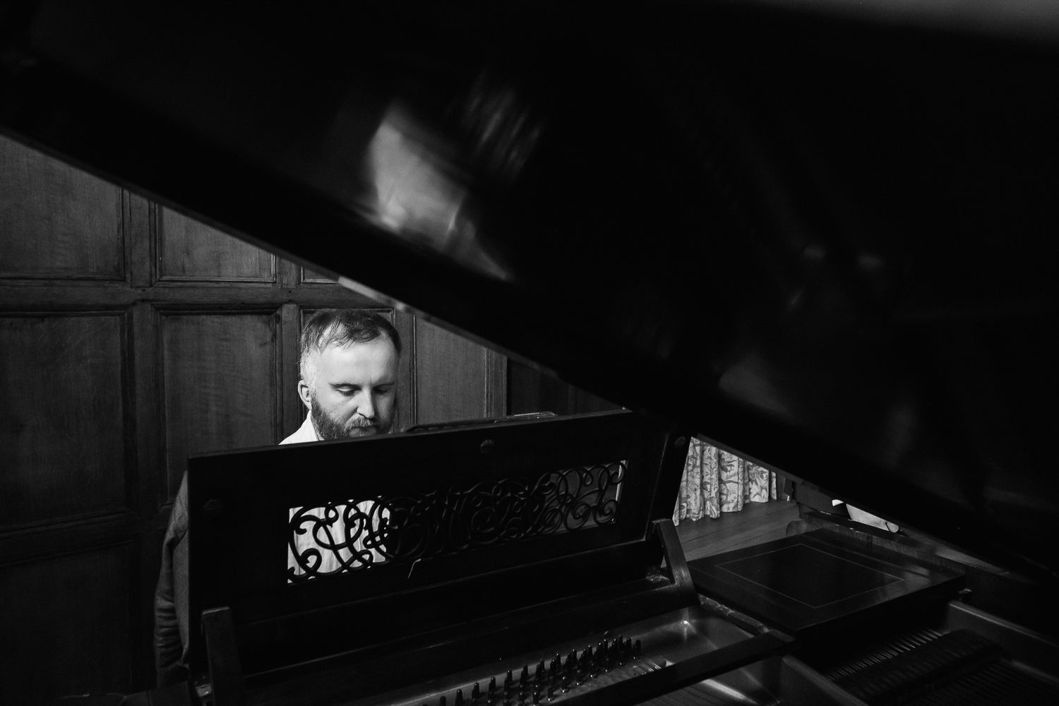 Black and white photo of a man playing a grand piano in a dimly lit room with wood-paneled walls.