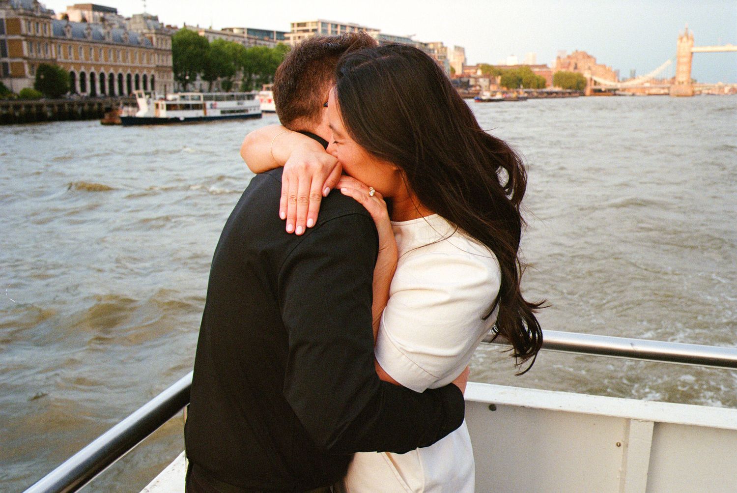 Couple embracing on boat during sunset with London cityscape in background.