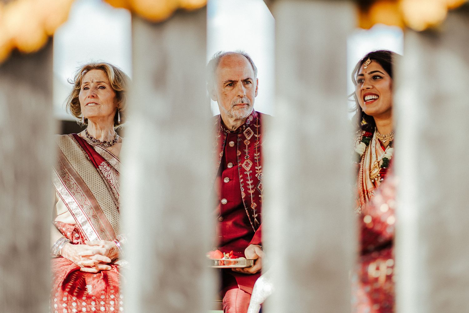 Three people in formal traditional attire seen through decorative wedding ceremony elements.