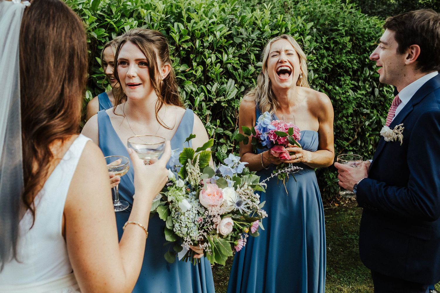 Wedding party members sharing laughs and holding bouquets in a garden setting.