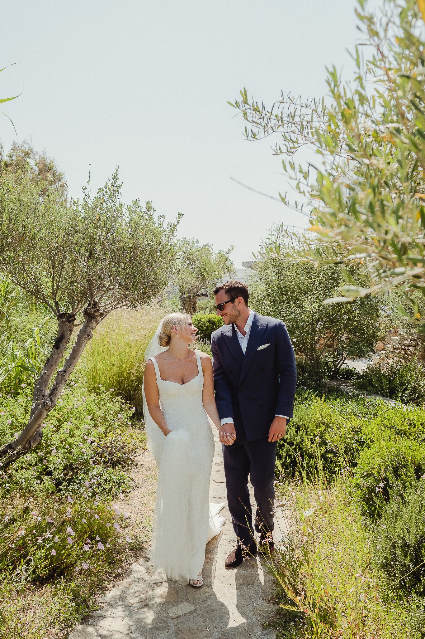 A couple walking together on a rustic path surrounded by olive trees and lush greenery in a Mediterranean setting.