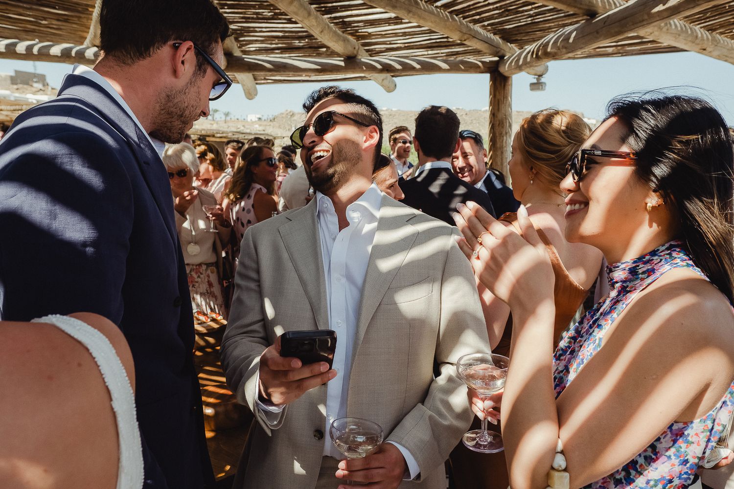 Wedding guests gather under a rustic wooden pergola for an outdoor celebration.