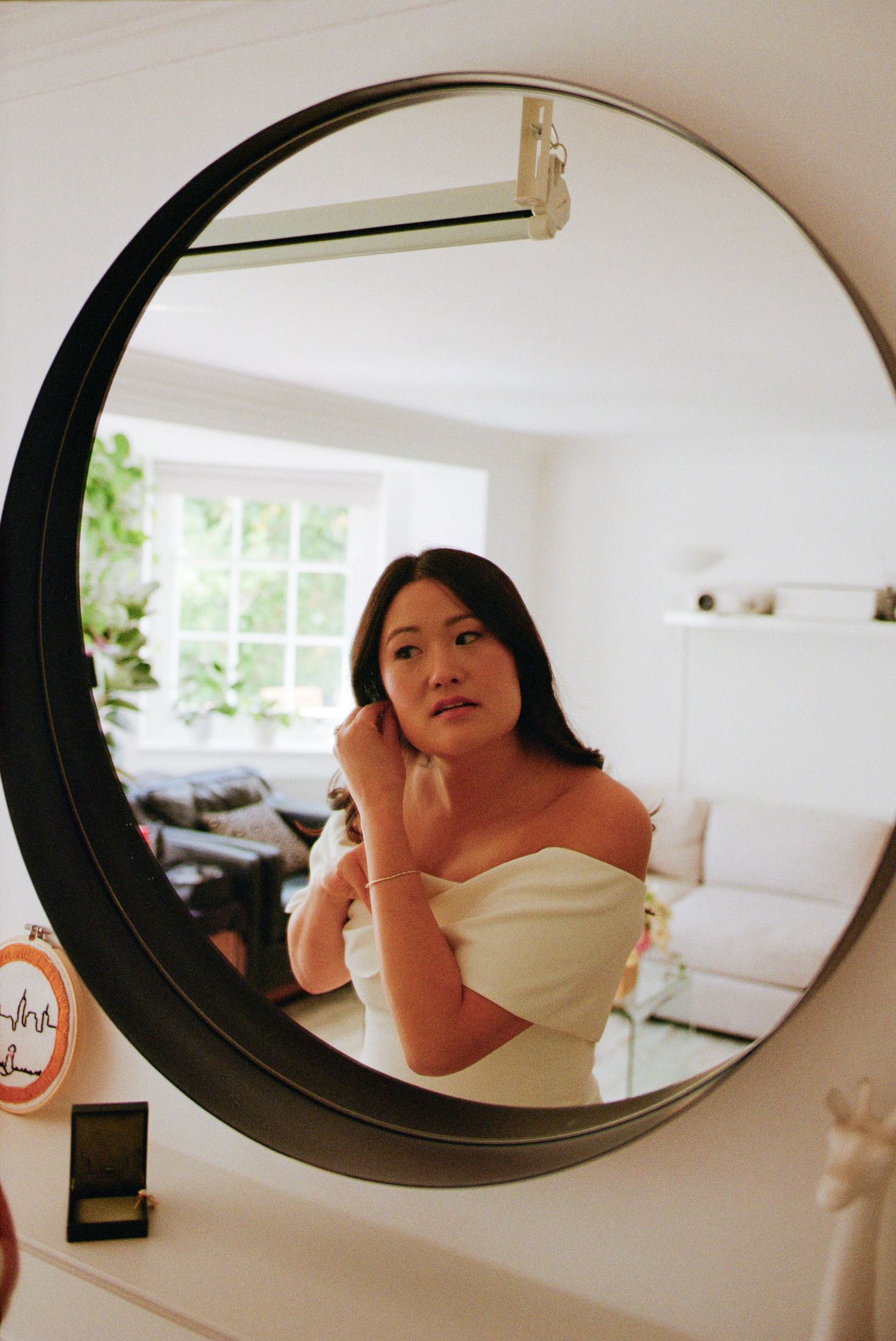 Bride in white off-shoulder gown adjusting earring while looking into a round black-framed mirror.