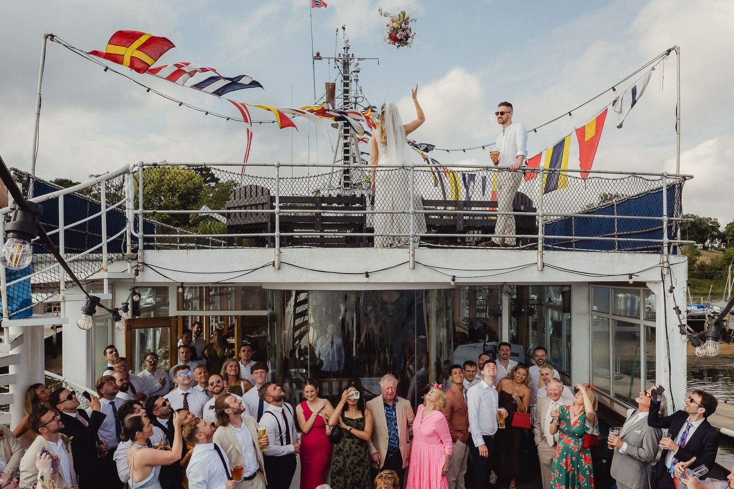 Large crowd gathered on a boat deck with colorful flags and banners flying overhead during a maritime celebration.