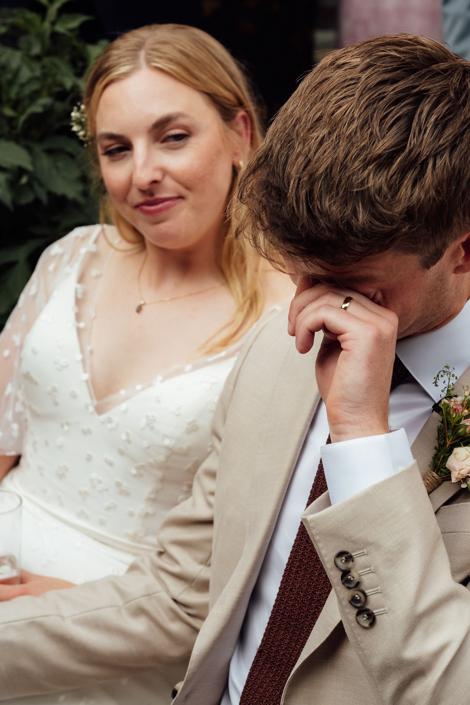 A smiling bride in a white lace wedding dress stands beside a groom in a beige suit who wipes his eye with his hand.
