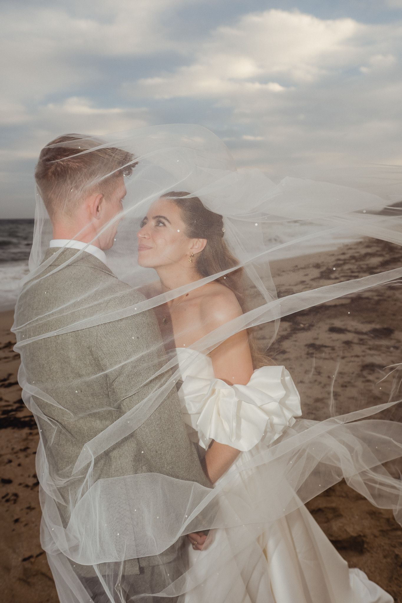 A flowing white wedding veil billows dramatically in the wind against a cloudy coastal backdrop.