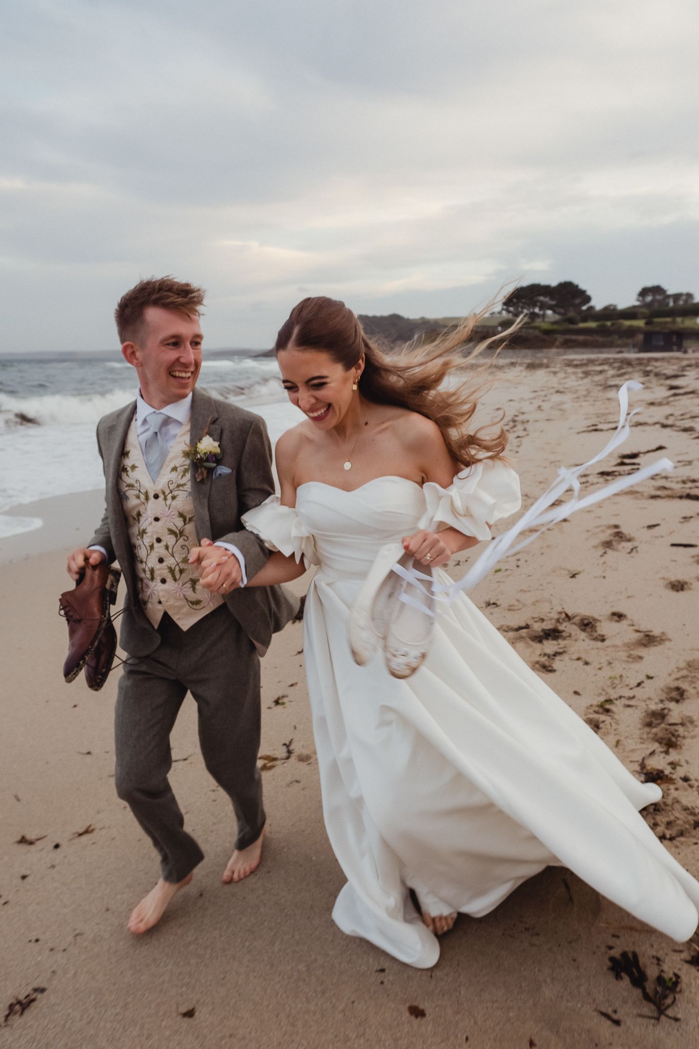 A couple runs and laughs together on a windy beach during their wedding photoshoot, with the bride's dress flowing in the breeze.