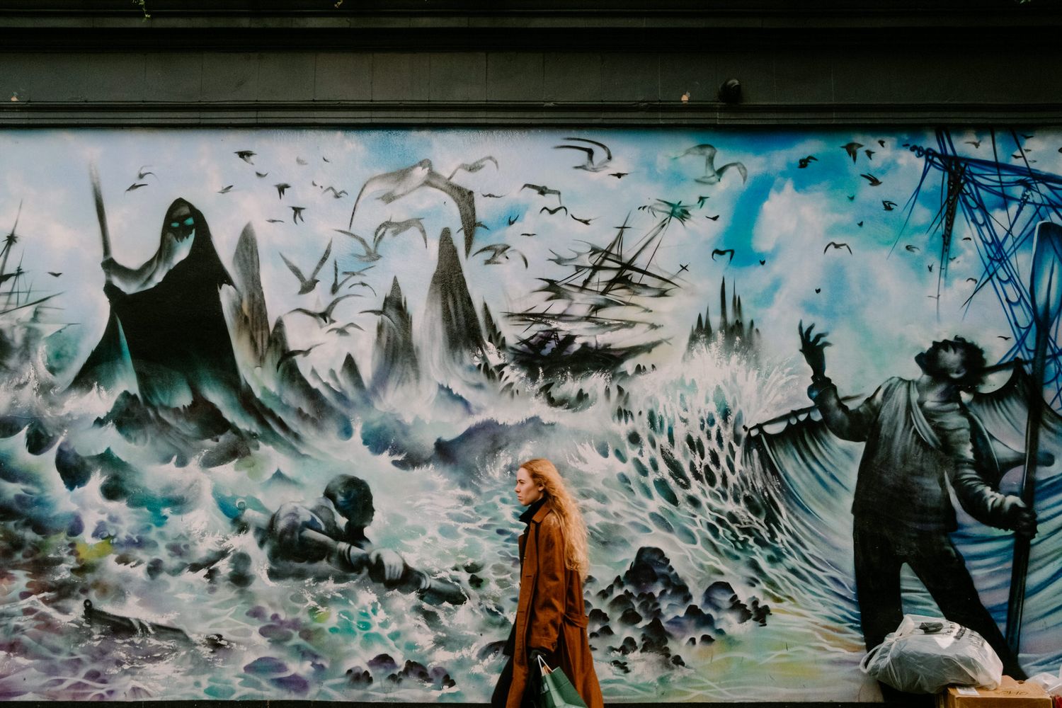 A person in an orange coat walks past a dramatic street art mural depicting a stormy ocean scene in black and turquoise colors.