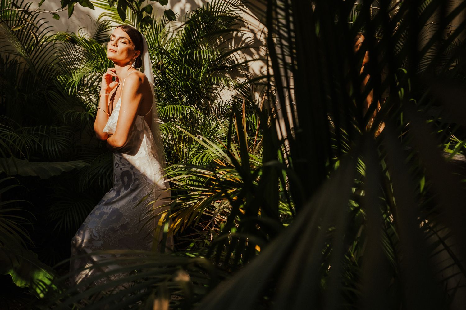 Bride in fitted white dress stands among lush palm fronds in dappled sunlight.