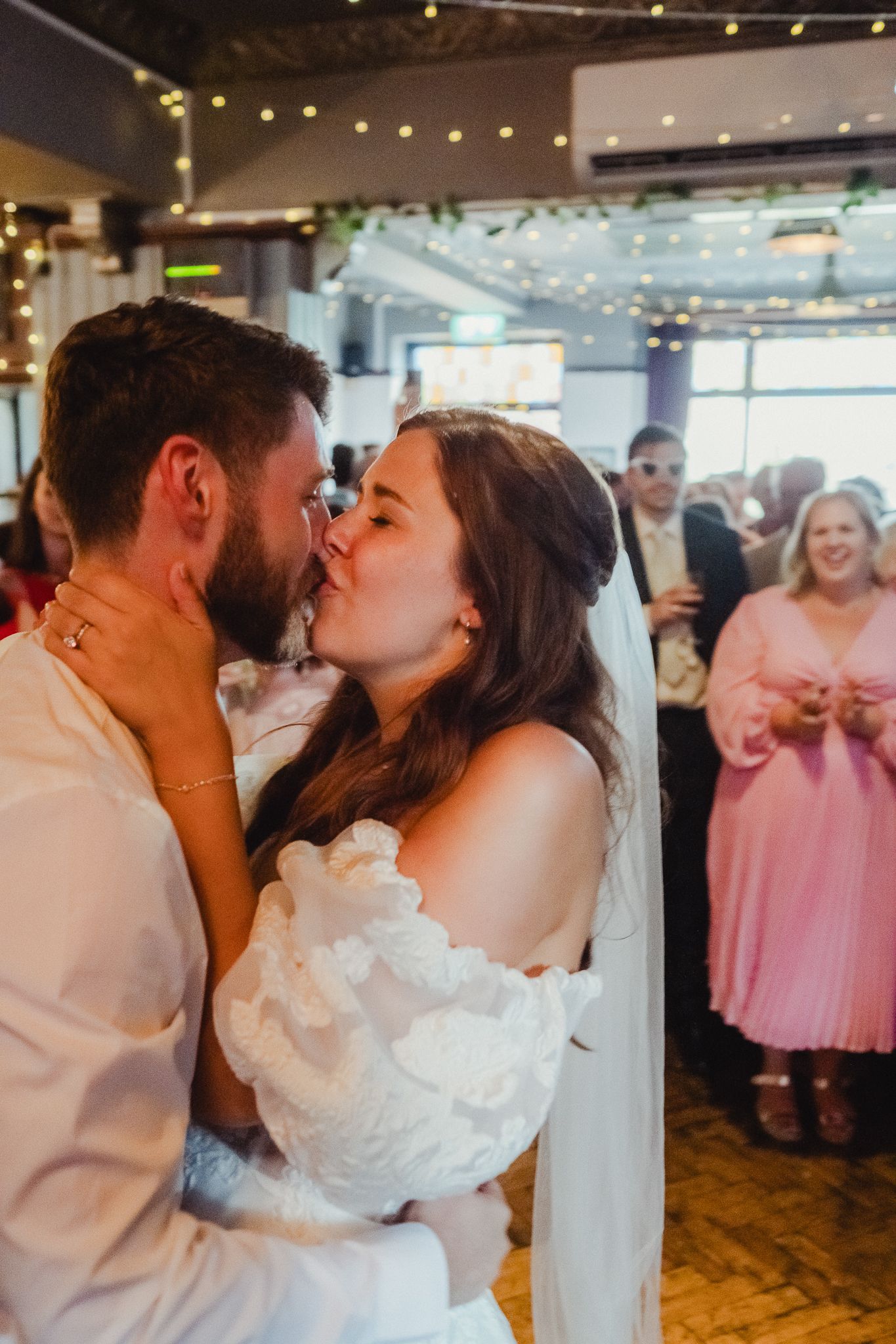 Romantic wedding couple shares their first dance kiss while guests watch in a beautifully lit reception venue.