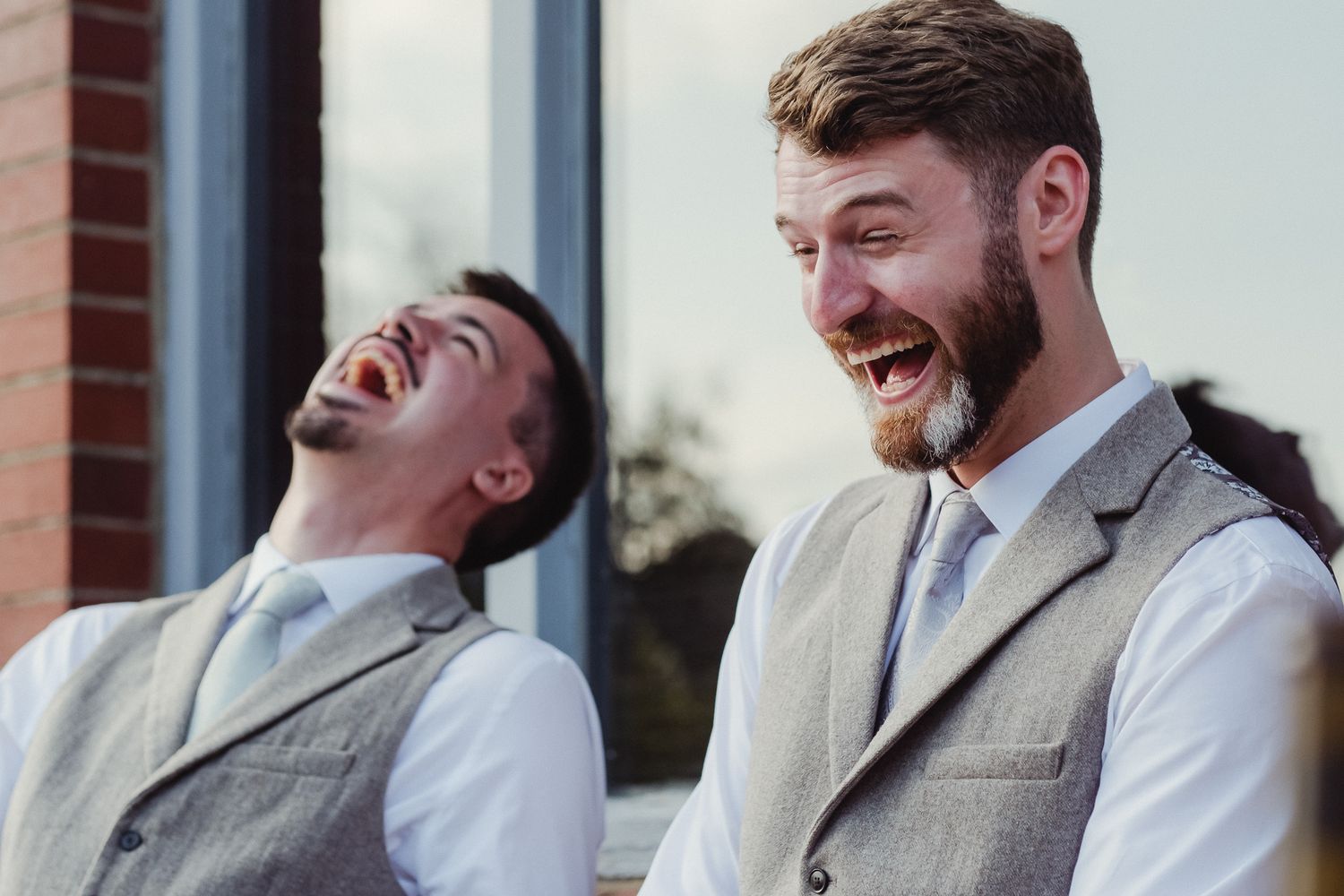Two people in matching gray vests and suits share a candid moment of laughter outdoors.