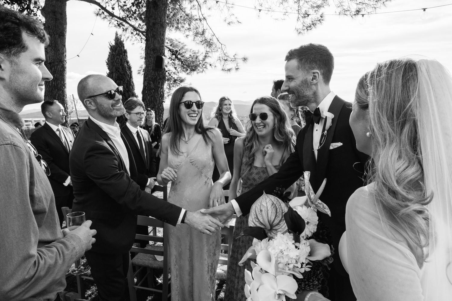 Black and white photo of wedding guests greeting a groom in a suit who shakes hands with a man as a bride holding flowers stands nearby.