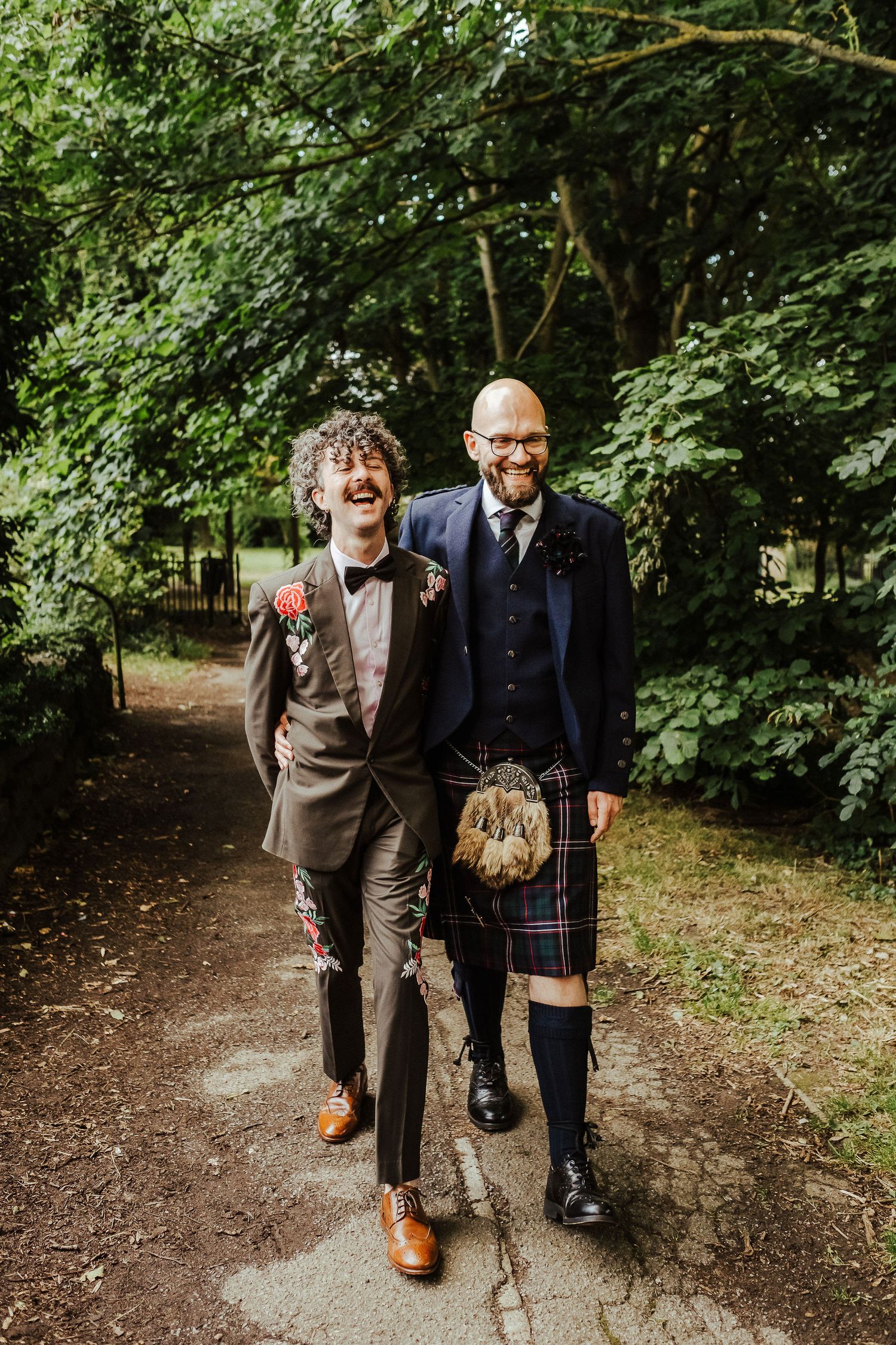 Two grooms walk arm in arm along a leafy garden path, one in a brown suit with a floral boutonniere, the other in a navy jacket and tartan k