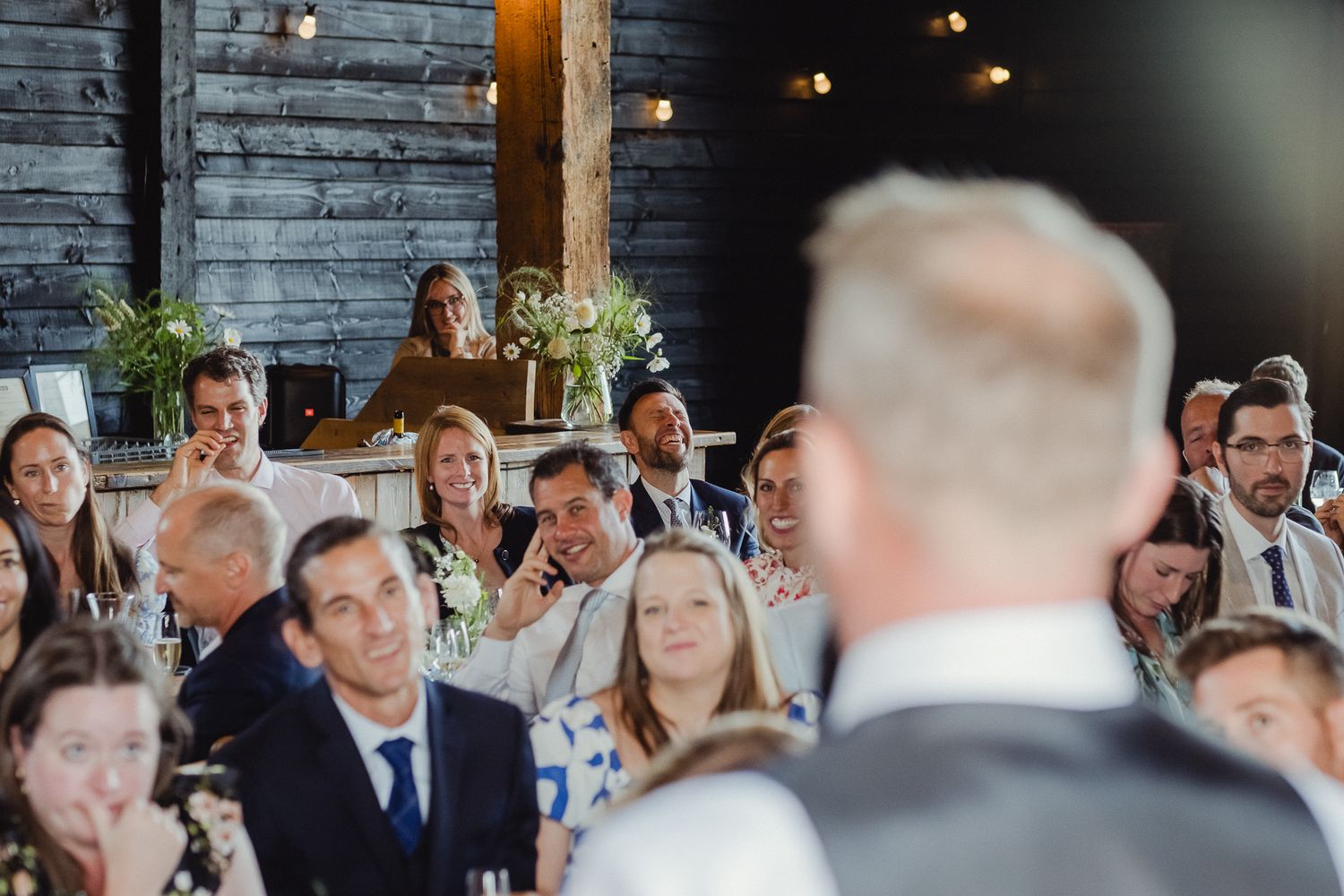 Wedding reception guests seated at long tables in a rustic barn with wooden beams and warm lighting.