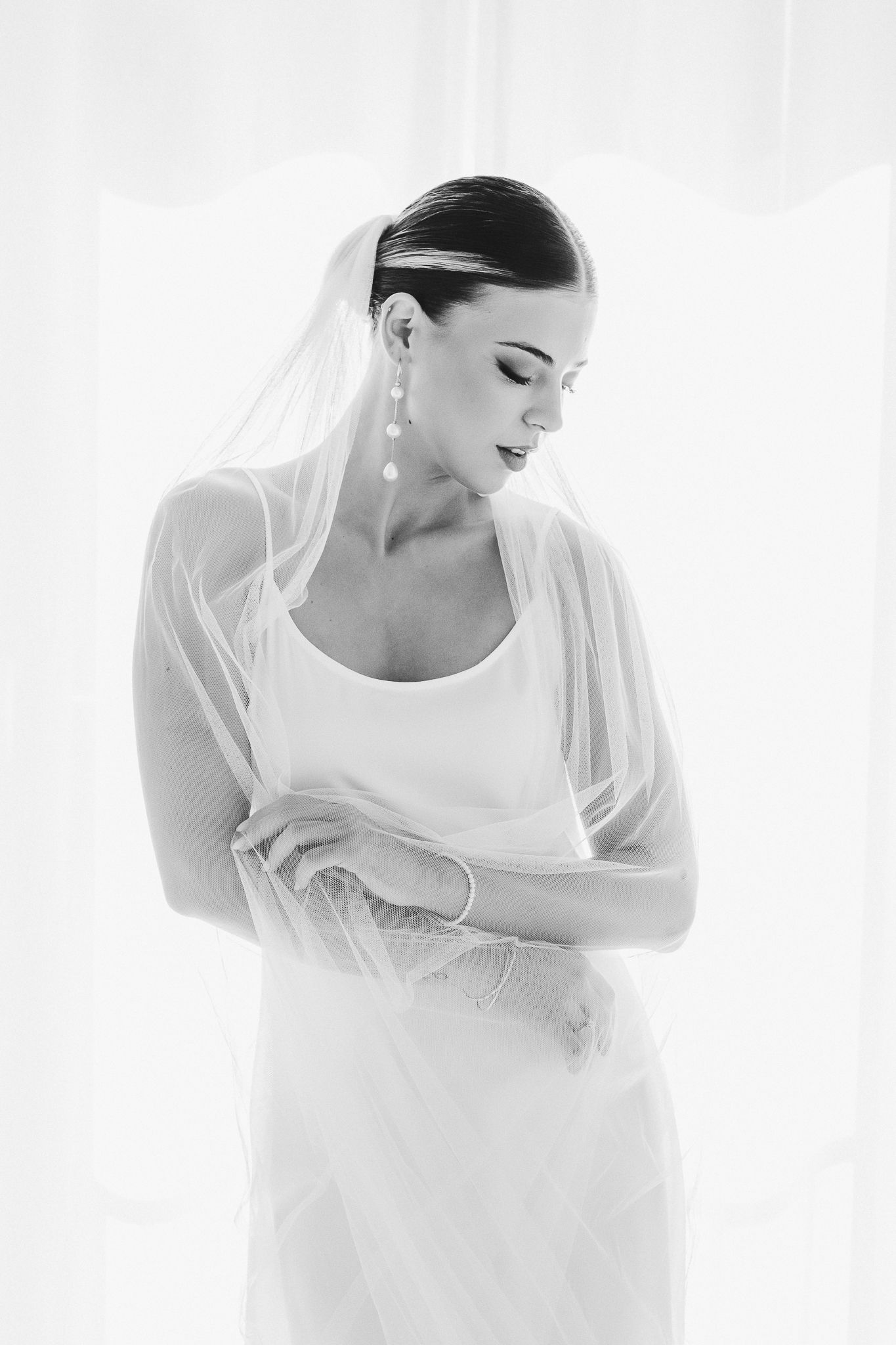 Artistic black and white portrait of bride in flowing white dress and veil against bright window light.