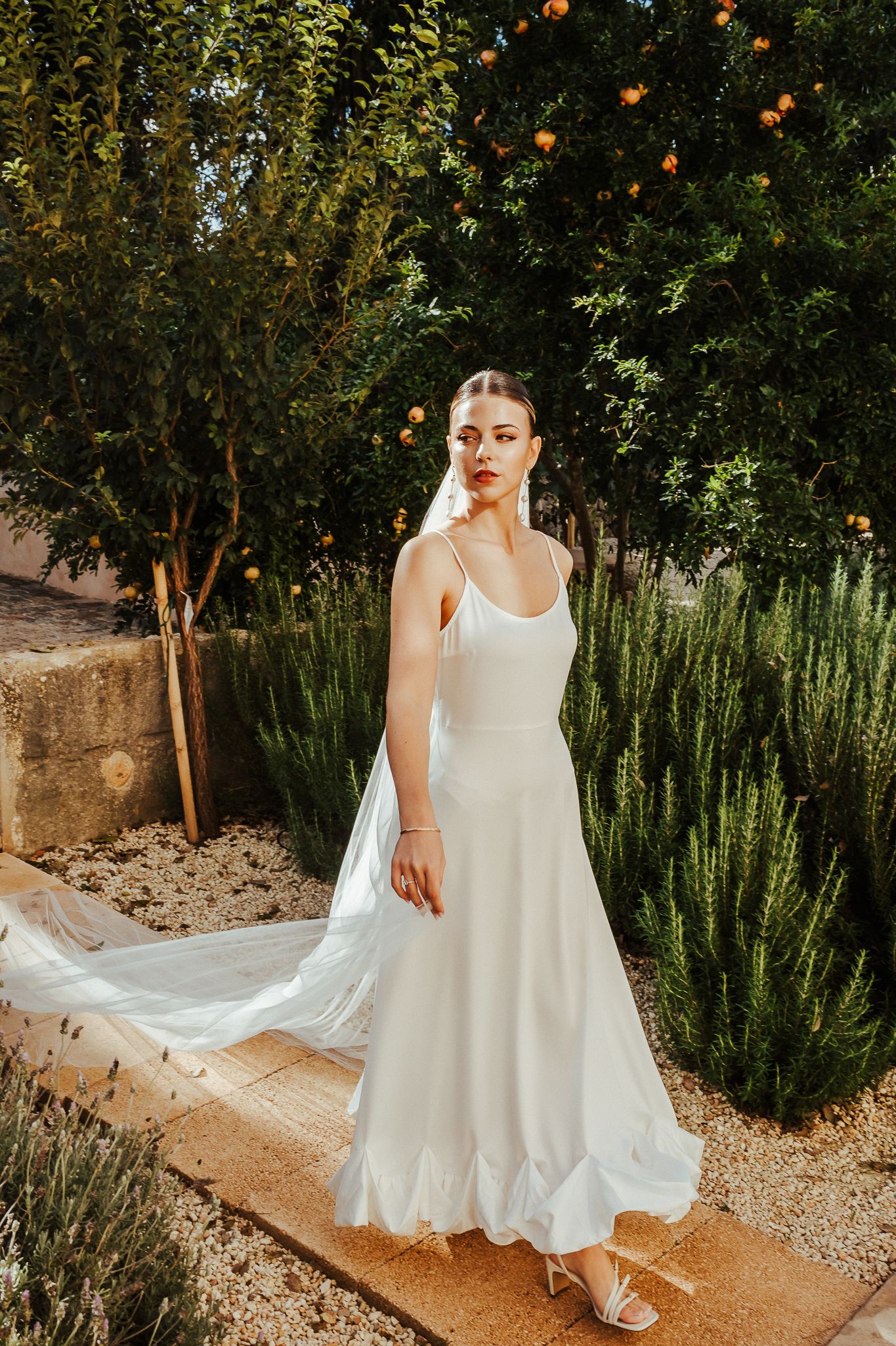 A flowing white dress twirls gracefully along a garden path lined with citrus trees and rosemary bushes.