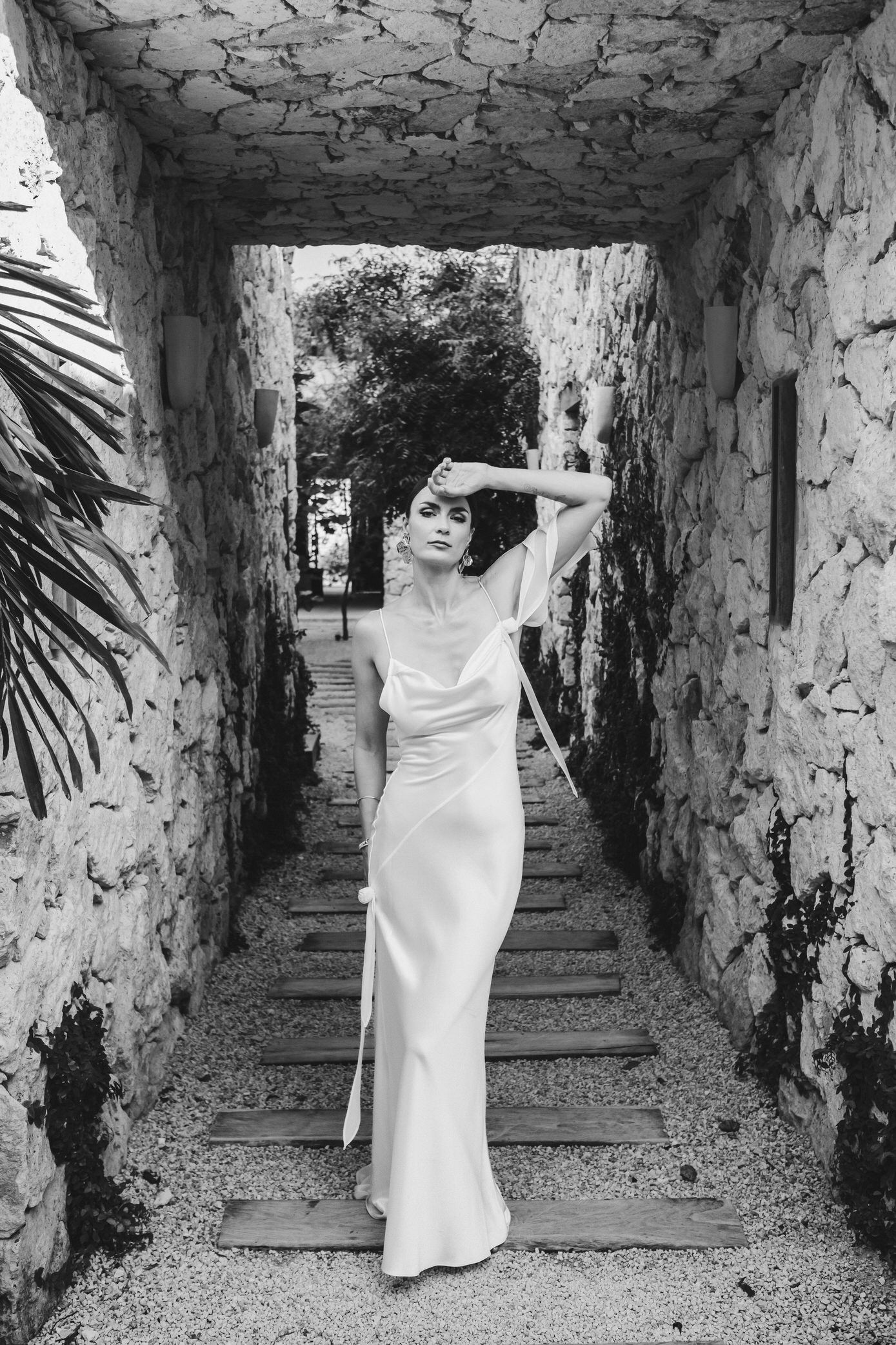 Elegant figure in flowing white dress poses gracefully in rustic stone archway corridor.