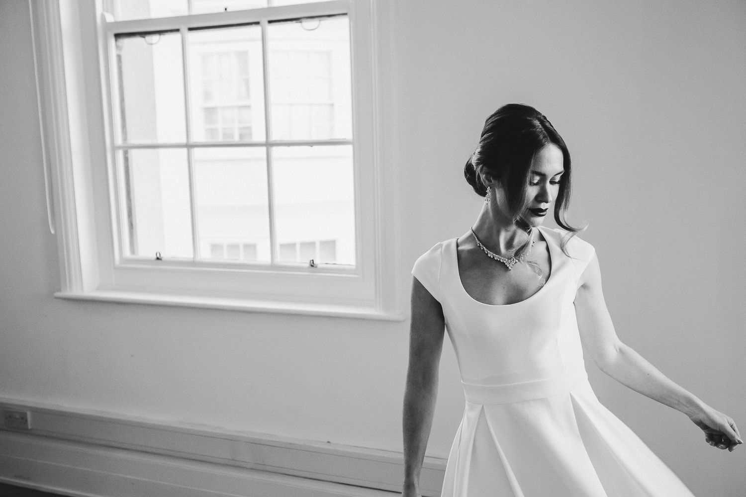 Black and white photo of someone in a white dress standing by a window in a bright room.
