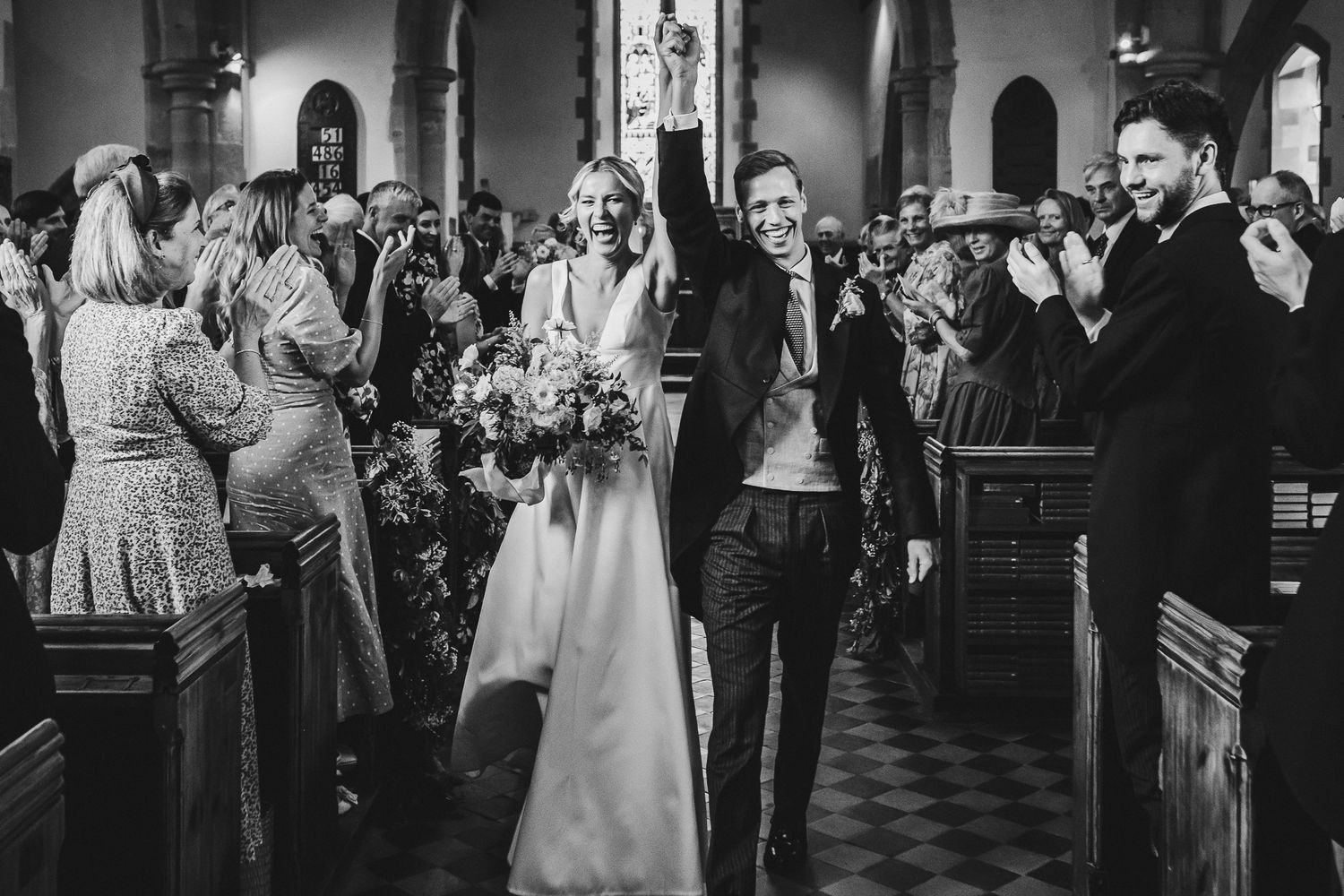 Wedding party exits church in celebration while guests applaud along the aisle.