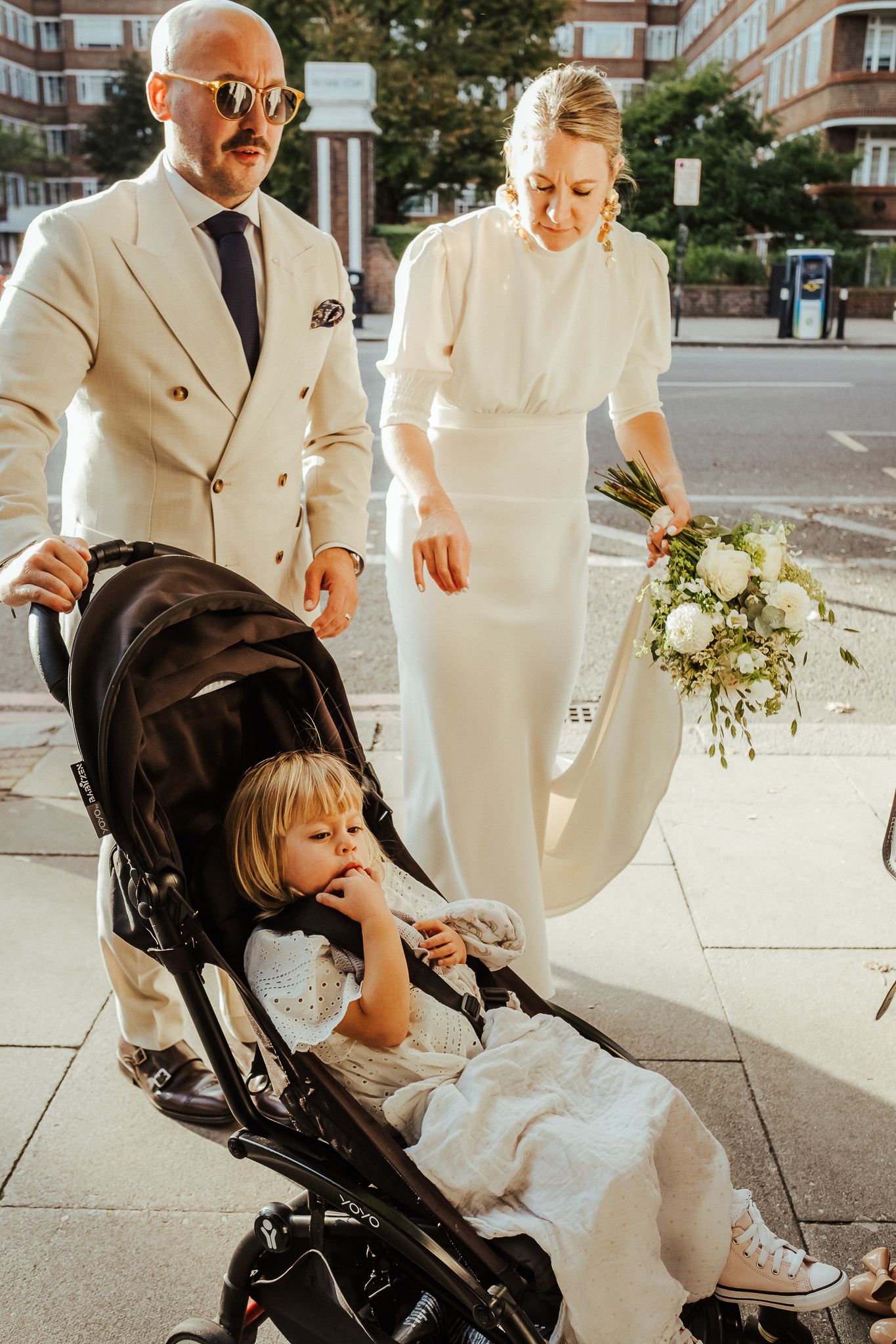 Two people walk down a city street in formal attire while a child sits in a stroller.